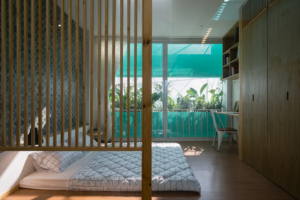 A minimalist bedroom featuring a floor mattress behind a vertical wood slat partition, next to a wooden wardrobe and balcony window.