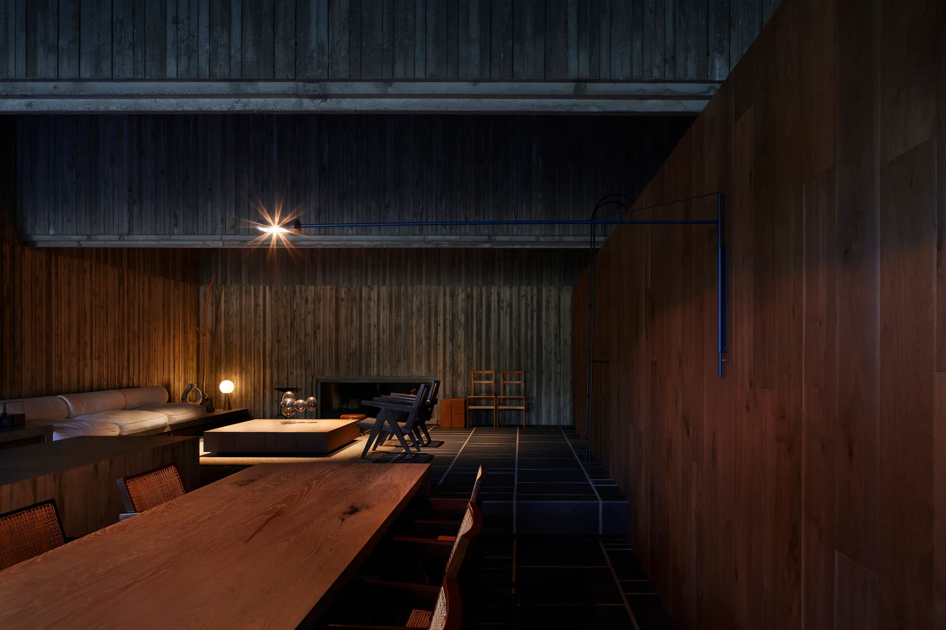 A moody open-plan living and dining space featuring vertical wood-slat walls, a large timber table, a low-profile white sofa, and dark stone floors.