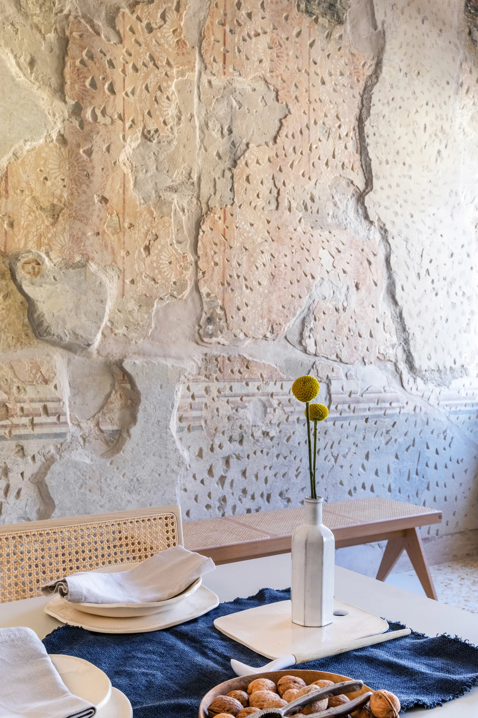 A dining table features ceramic dishware and a navy blue runner in front of a rustic, heavily distressed plaster wall and cane seating.