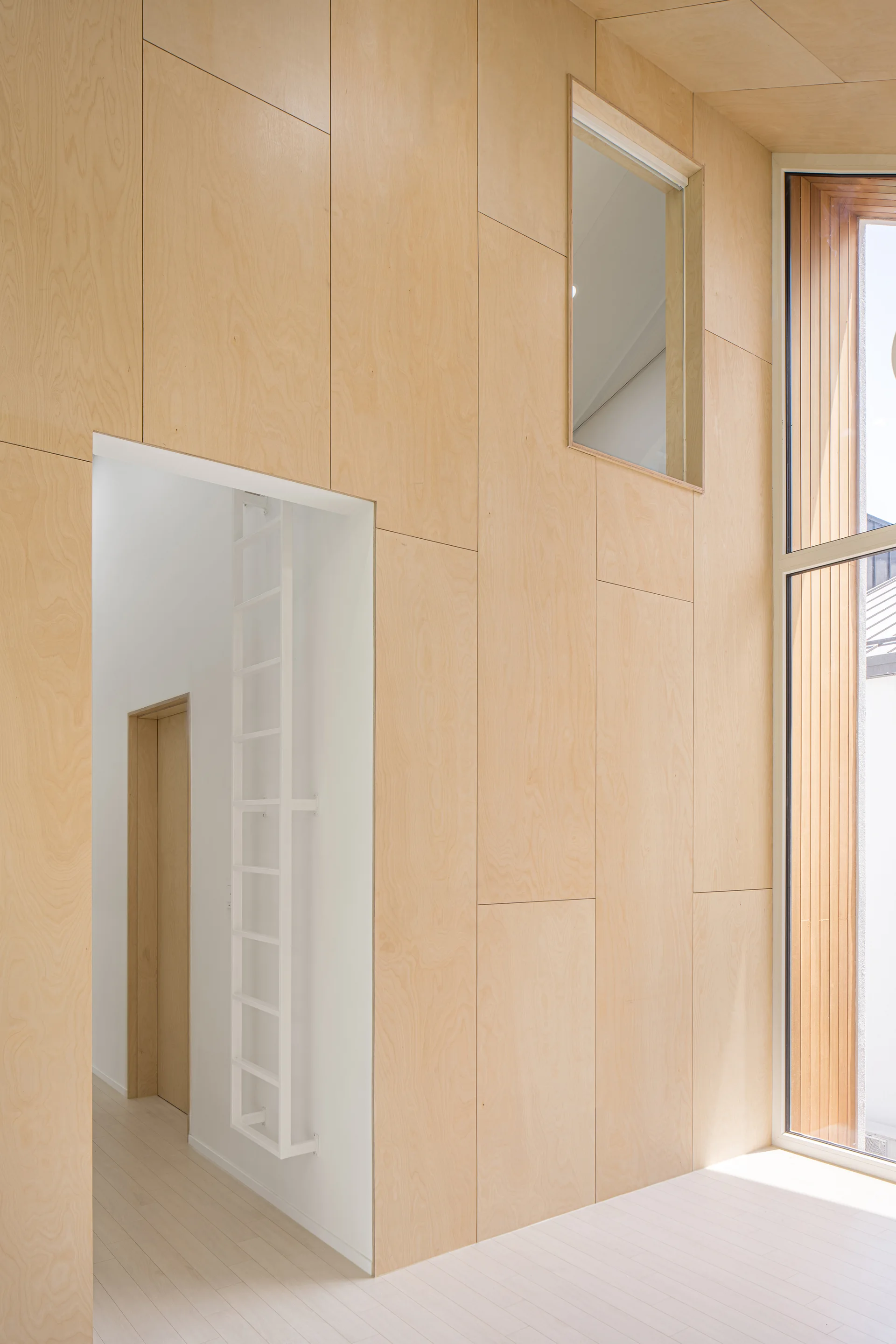 An interior room featuring light-colored plywood wall paneling, a white wall-mounted ladder inside an open doorway, and a tall window.
