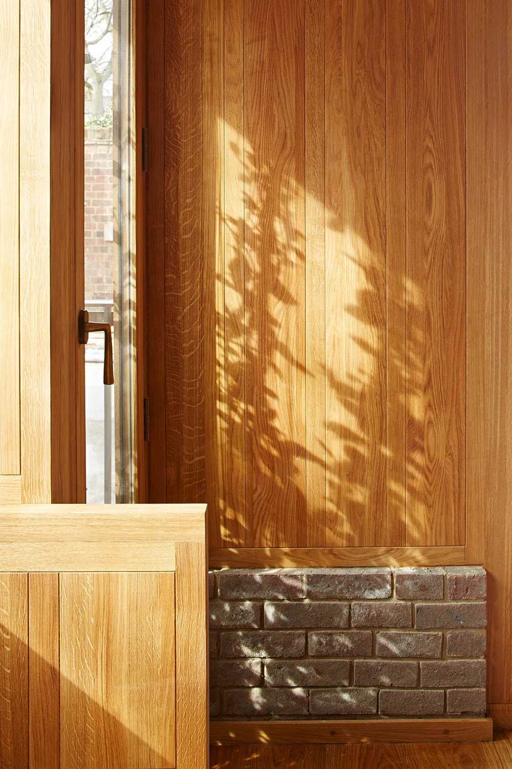 Vertical warm wood wall paneling and matching wood window frame above a small section of exposed brick base with dappled shadow patterns.