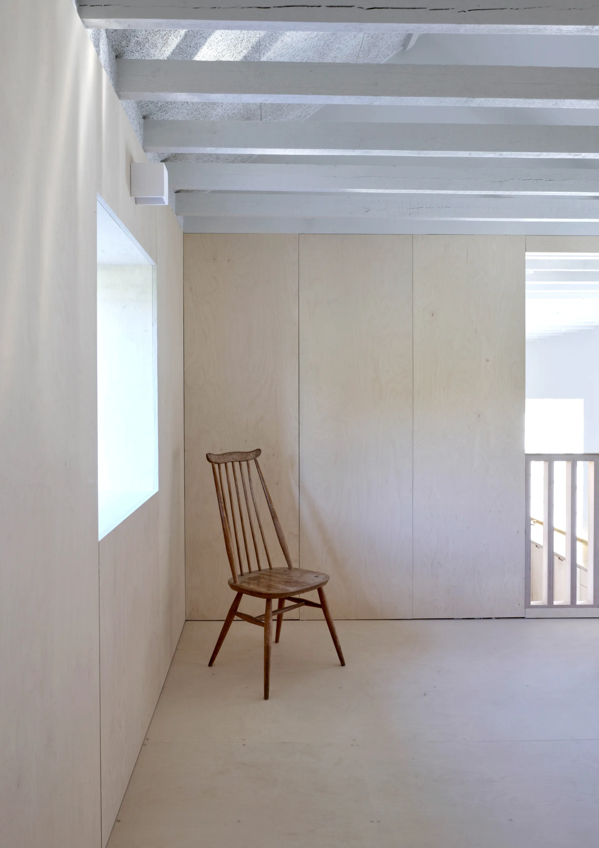 A minimalist landing features light plywood wall paneling, painted white ceiling beams, and a single wooden spindle-back chair near a small window.