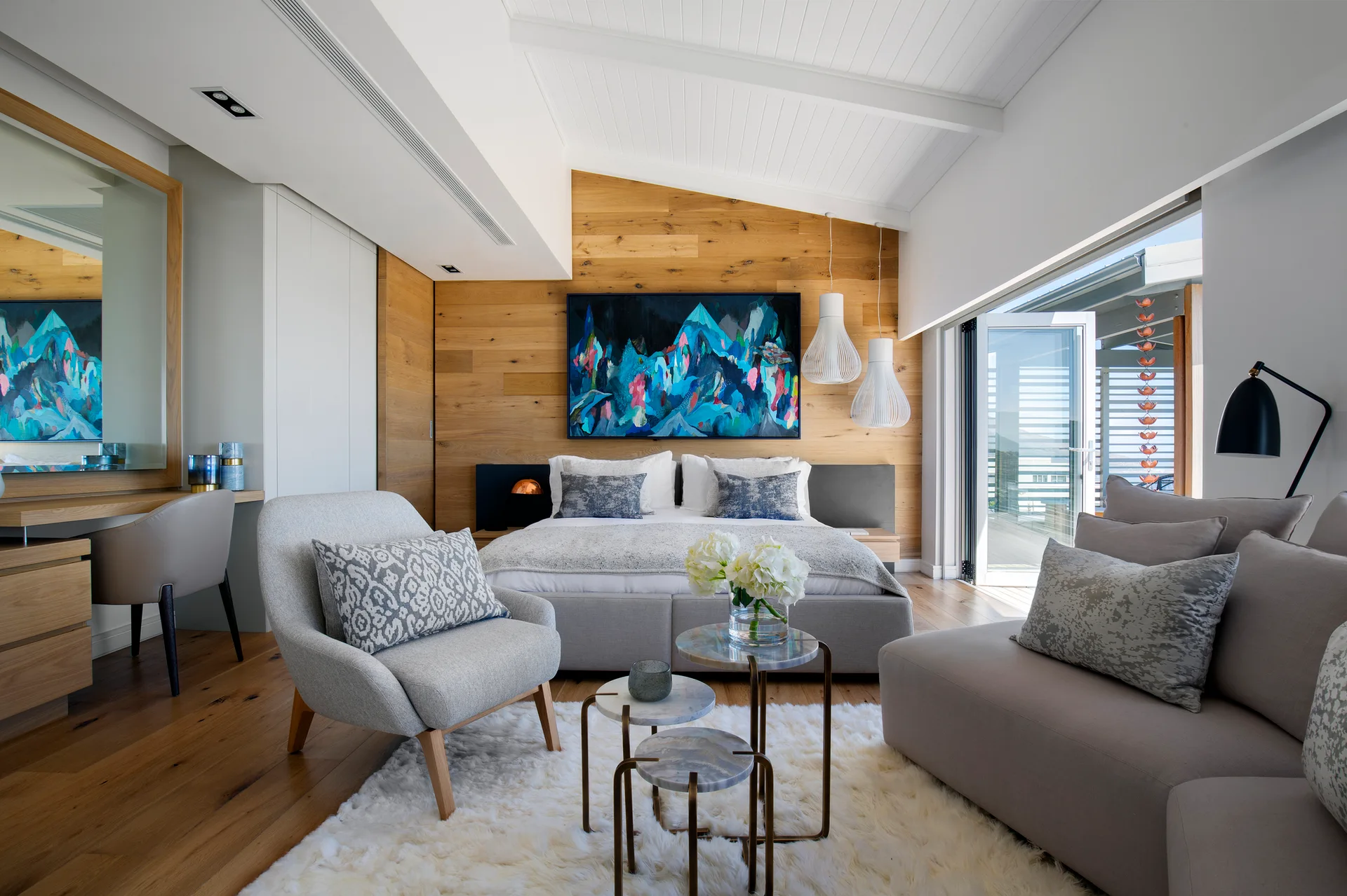 A modern bedroom features a wood paneled accent wall behind the bed, white vaulted ceilings, a gray armchair, and a plush white shag rug.