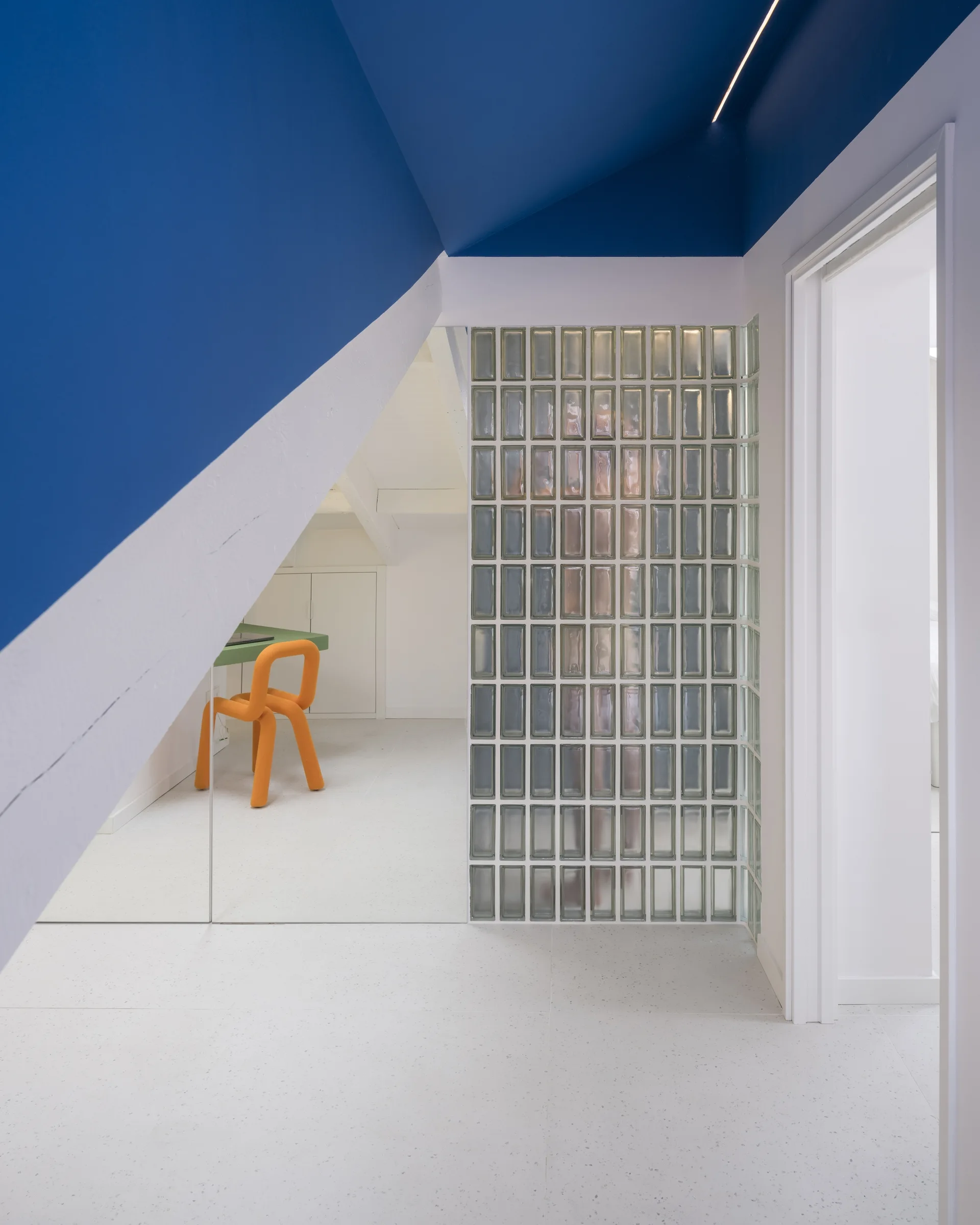 A modern home office features a bright blue sloped ceiling, a translucent glass block partition wall, white terrazzo-style floors, and a yellow chair.