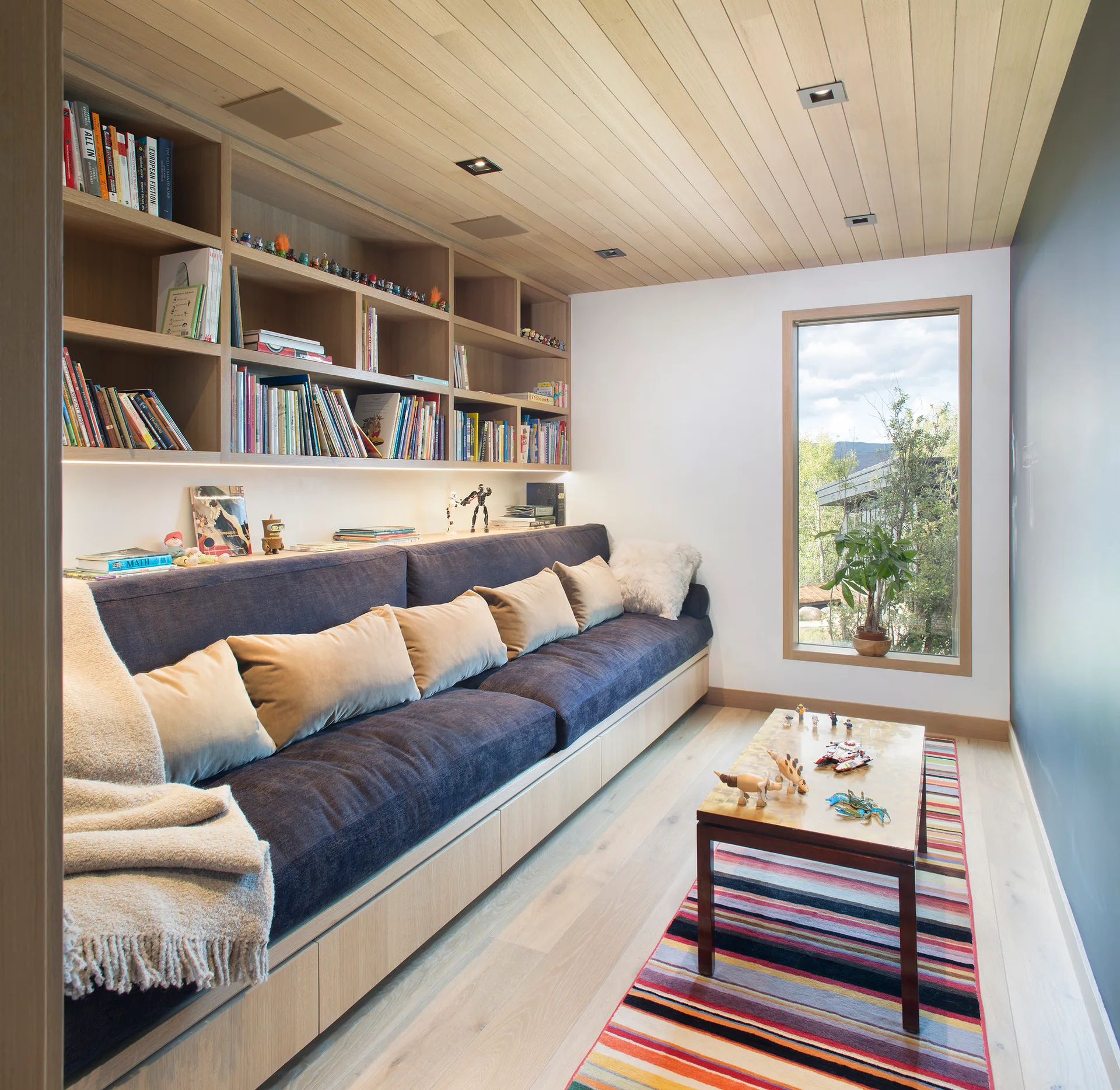 A narrow room features a custom light wood bookshelf with an integrated navy blue sofa, a wood-planked ceiling, and a colorful striped rug.