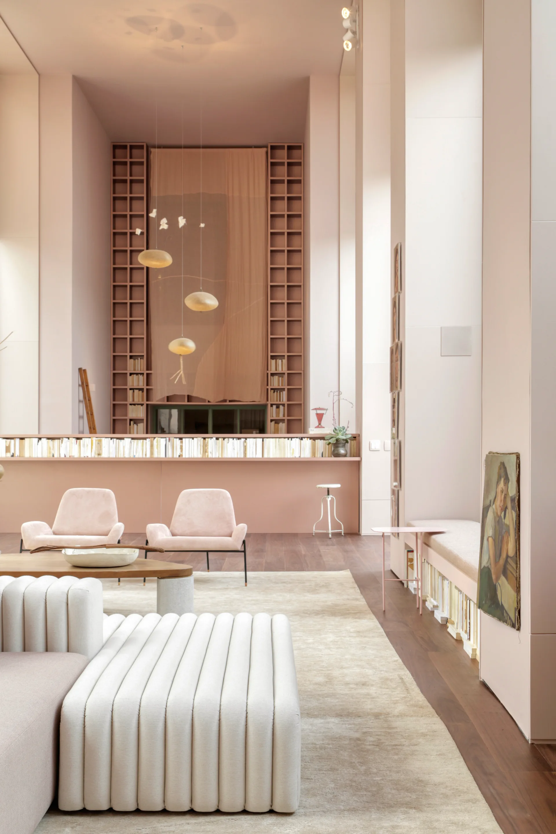 A double-height blush pink living room features a channel-tufted cream sofa, pale pink armchairs, and a tall grid-shelving unit in the background.