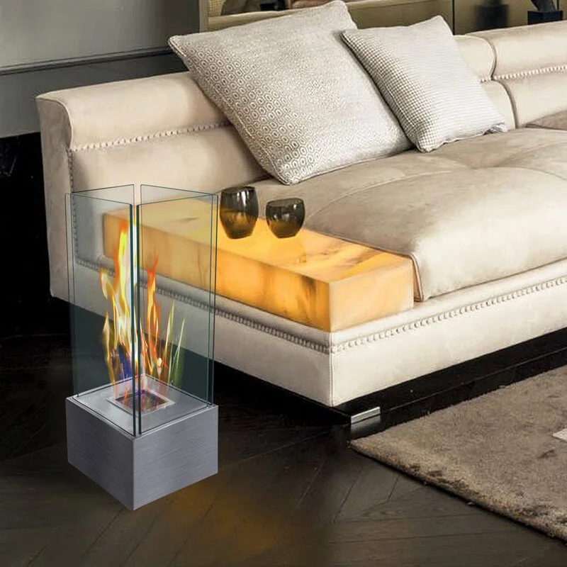 A light beige tufted sofa features an illuminated stone side table next to a small portable glass-enclosed fireplace on a dark herringbone wood floor.