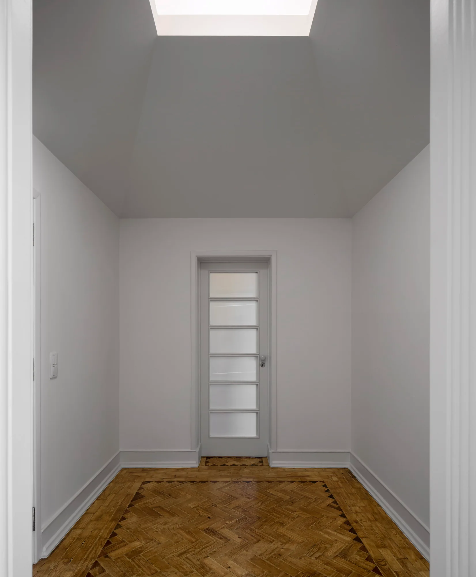 An empty white hallway with a glass pane door, herringbone parquet wood flooring, and a rectangular ceiling skylight.