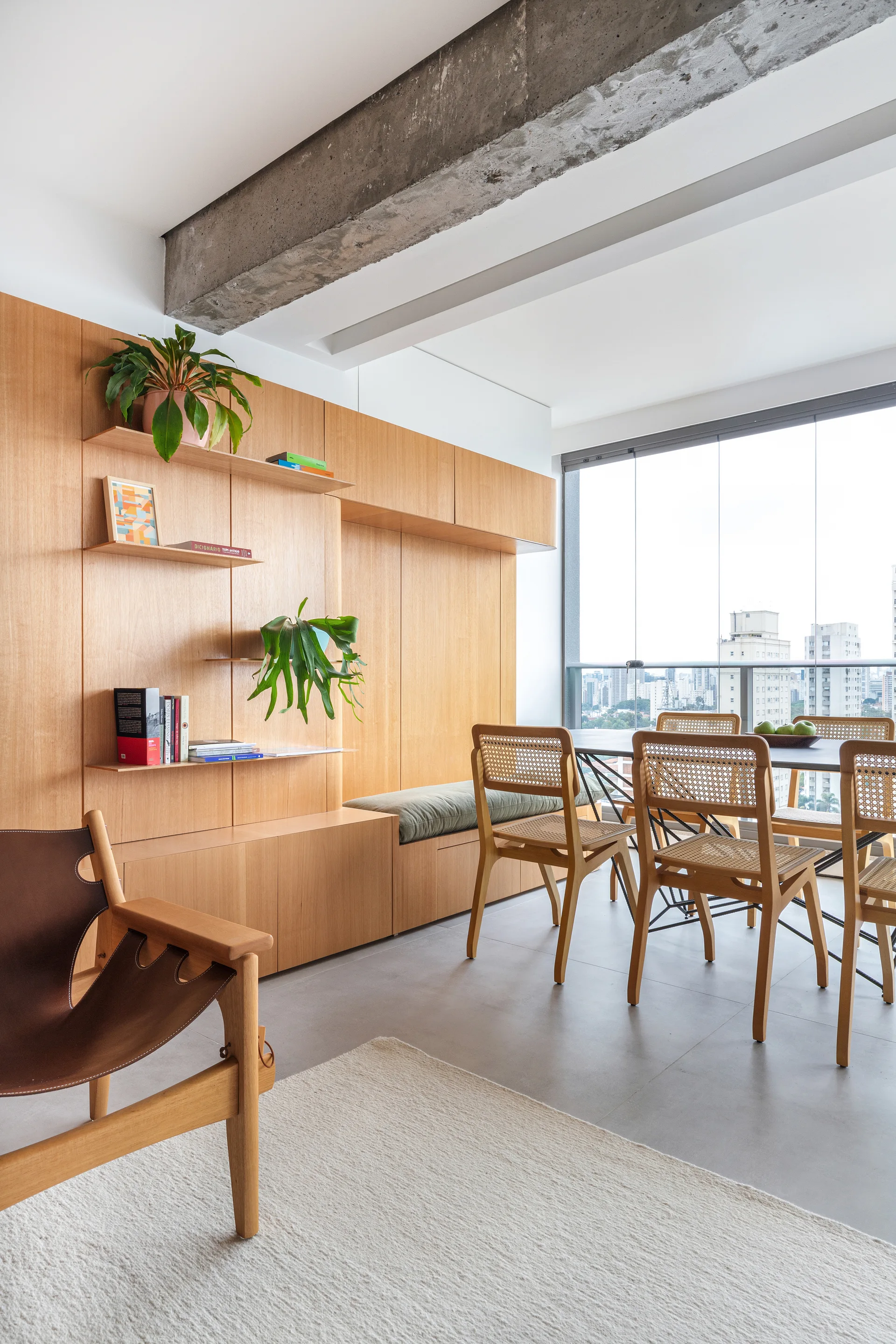 A modern dining room features a large wood-paneled storage wall, woven cane dining chairs around a table, and an exposed concrete ceiling beam.