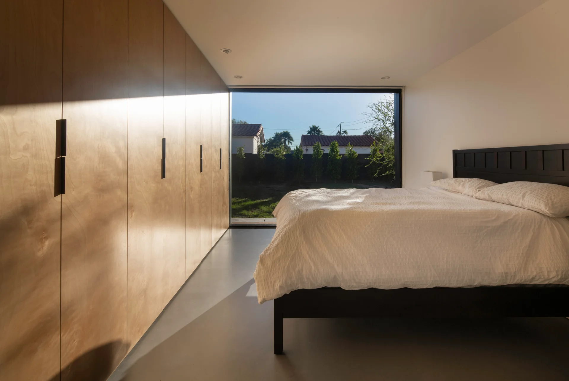 A minimalist bedroom featuring a floor-to-ceiling wood wardrobe, a black bed with white linens, and a large window facing an outdoor yard.