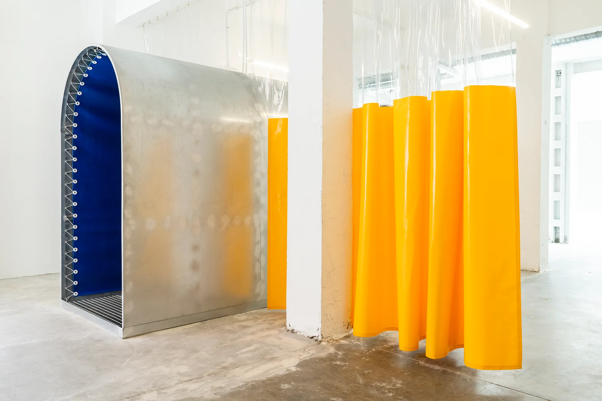 A brushed metal archway with a blue interior stands near a bright yellow hanging vinyl curtain partition in a minimalist white-walled space.