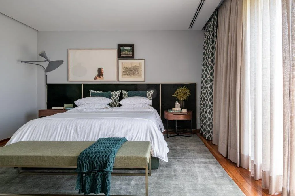 A contemporary bedroom features a bed with white bedding, a wide dark headboard panel, an upholstered green bench, and sheer curtains.