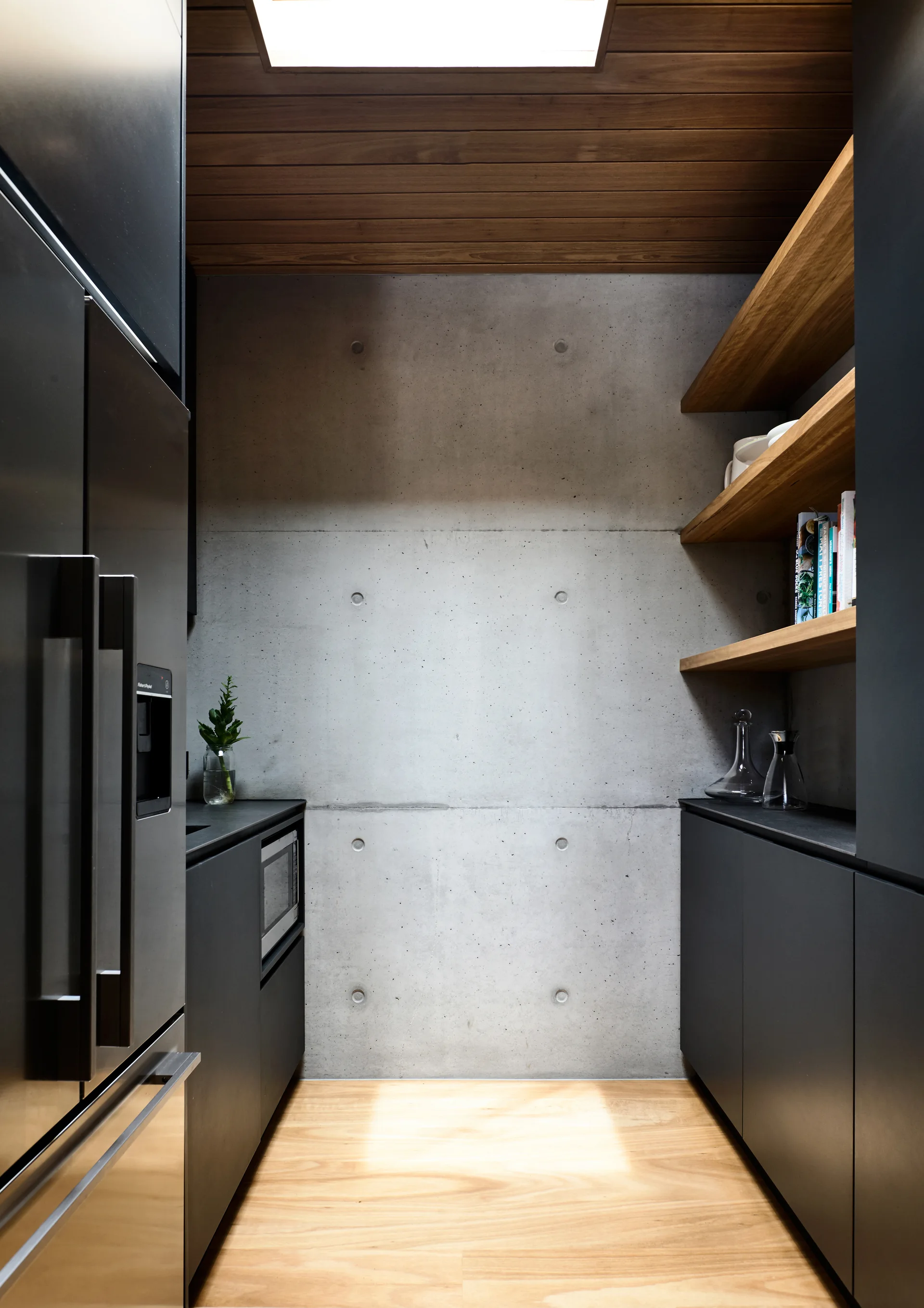 A modern narrow kitchen pantry features a concrete back wall, matte black cabinets, wooden open shelves, and a skylight-lit wood plank ceiling.