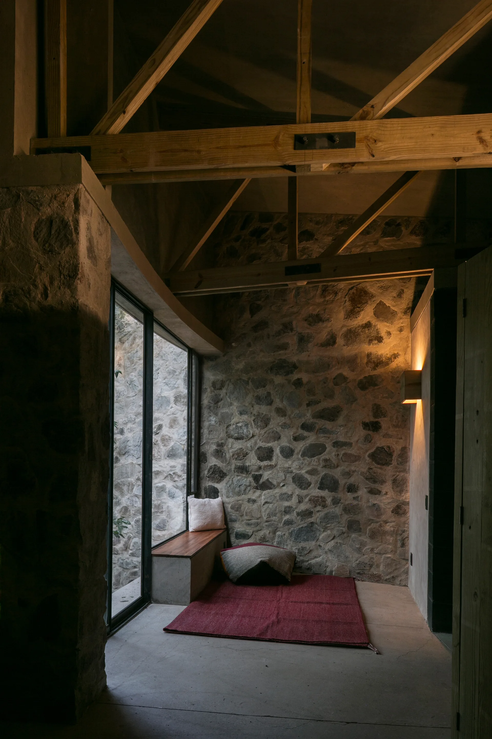 A rustic reading nook features exposed stone walls, timber ceiling trusses, a red floor rug, and a built-in window bench next to a tall glass window.