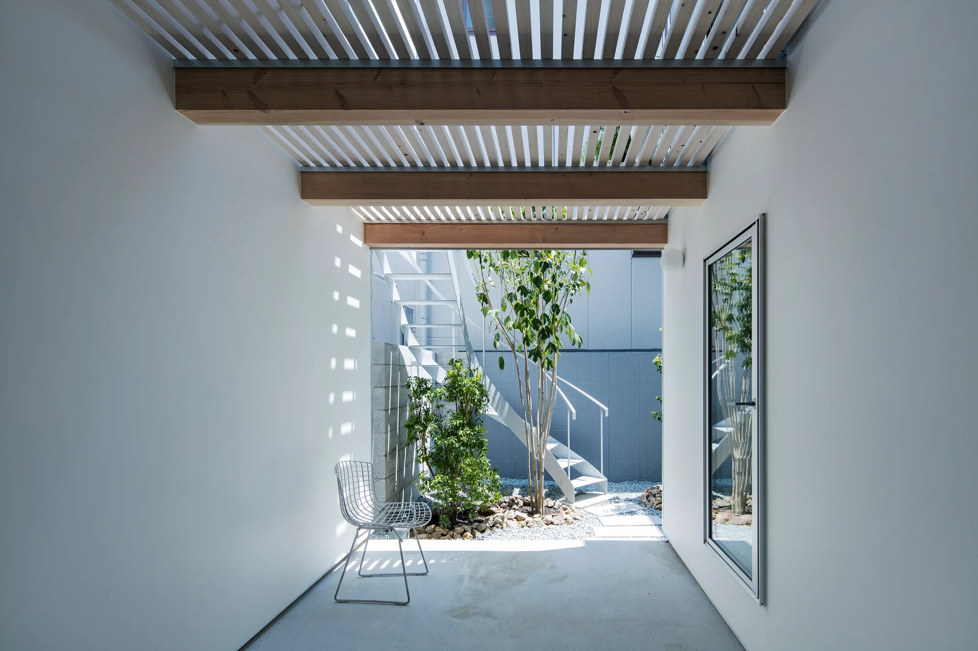 A minimalist white corridor with exposed wooden beams and a slatted ceiling leads to an open-air courtyard with a staircase and plants.