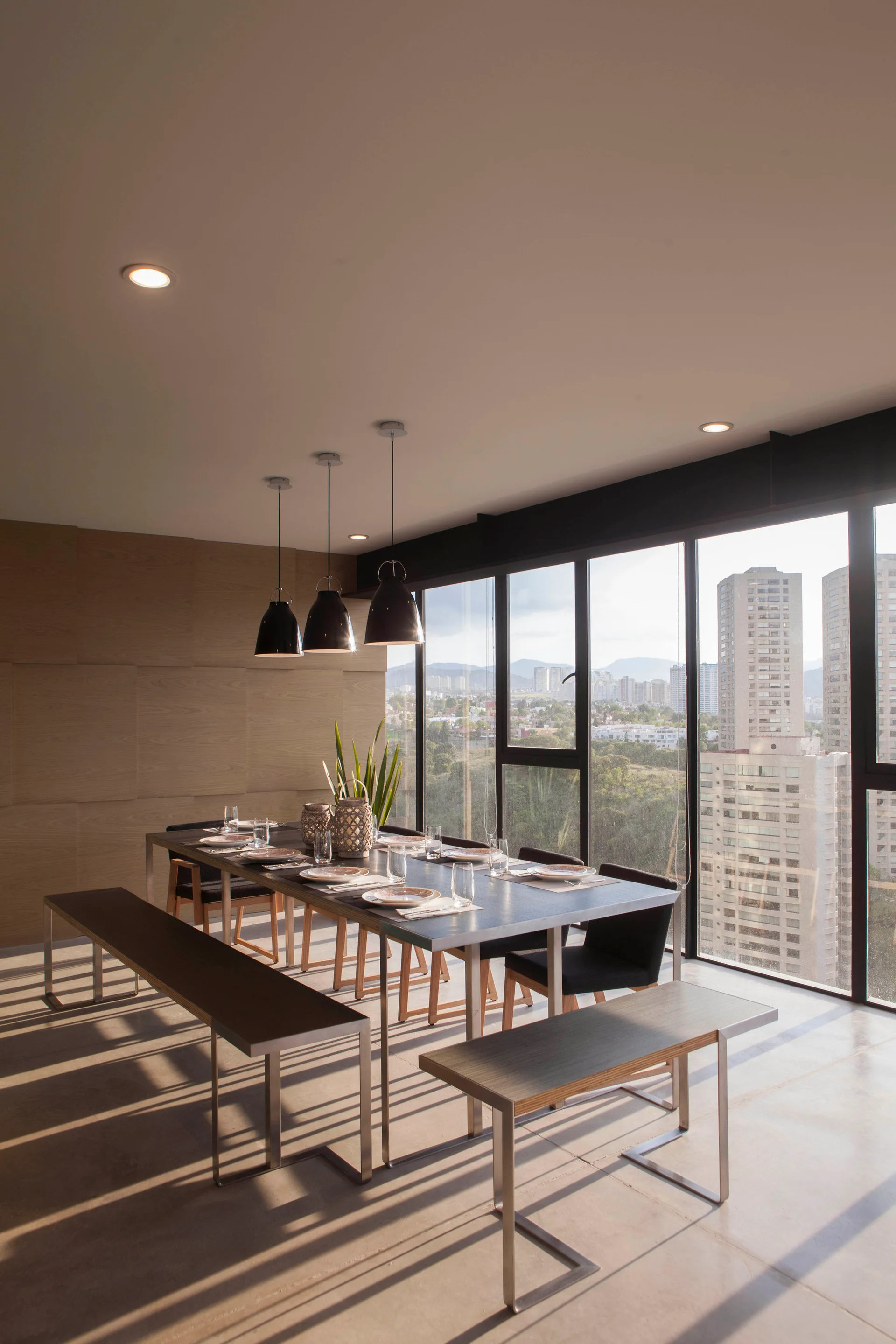 A minimalist dining room features a long table with matching wood benches, black pendant lights, a wood-paneled wall, and large windows showing a city view.