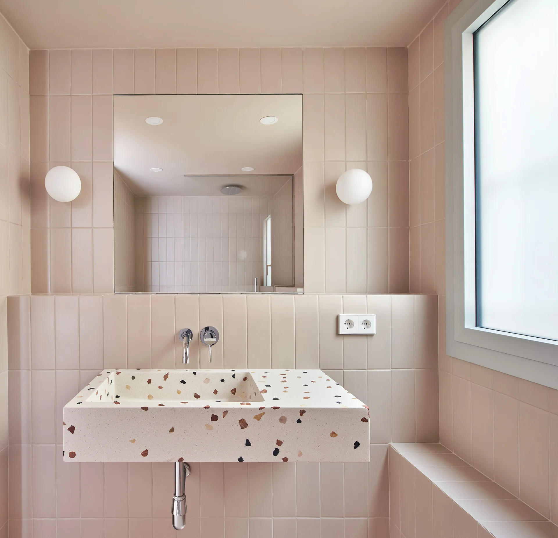 A minimalist bathroom features a floating terrazzo sink against light pink tiled walls, a square mirror, and two white globe sconces.