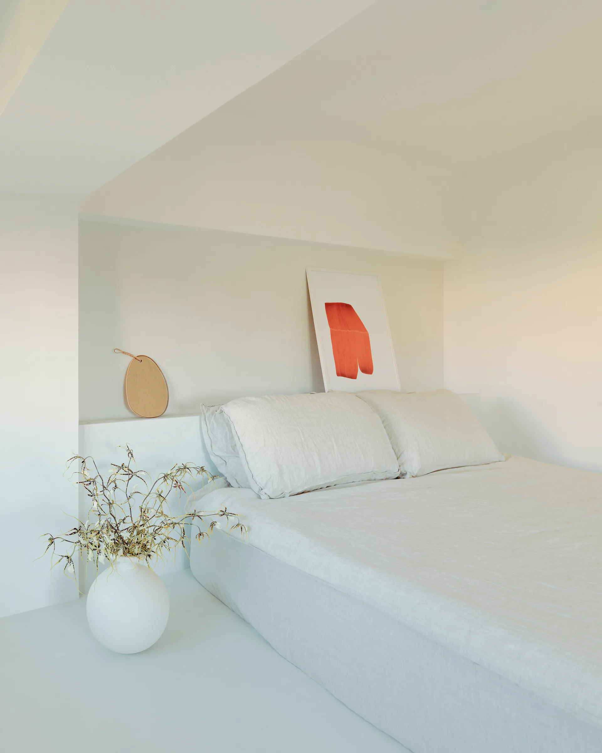 A minimalist bedroom featuring neutral linen bedding on a bed tucked into a plaster wall alcove next to a white ceramic floor vase.