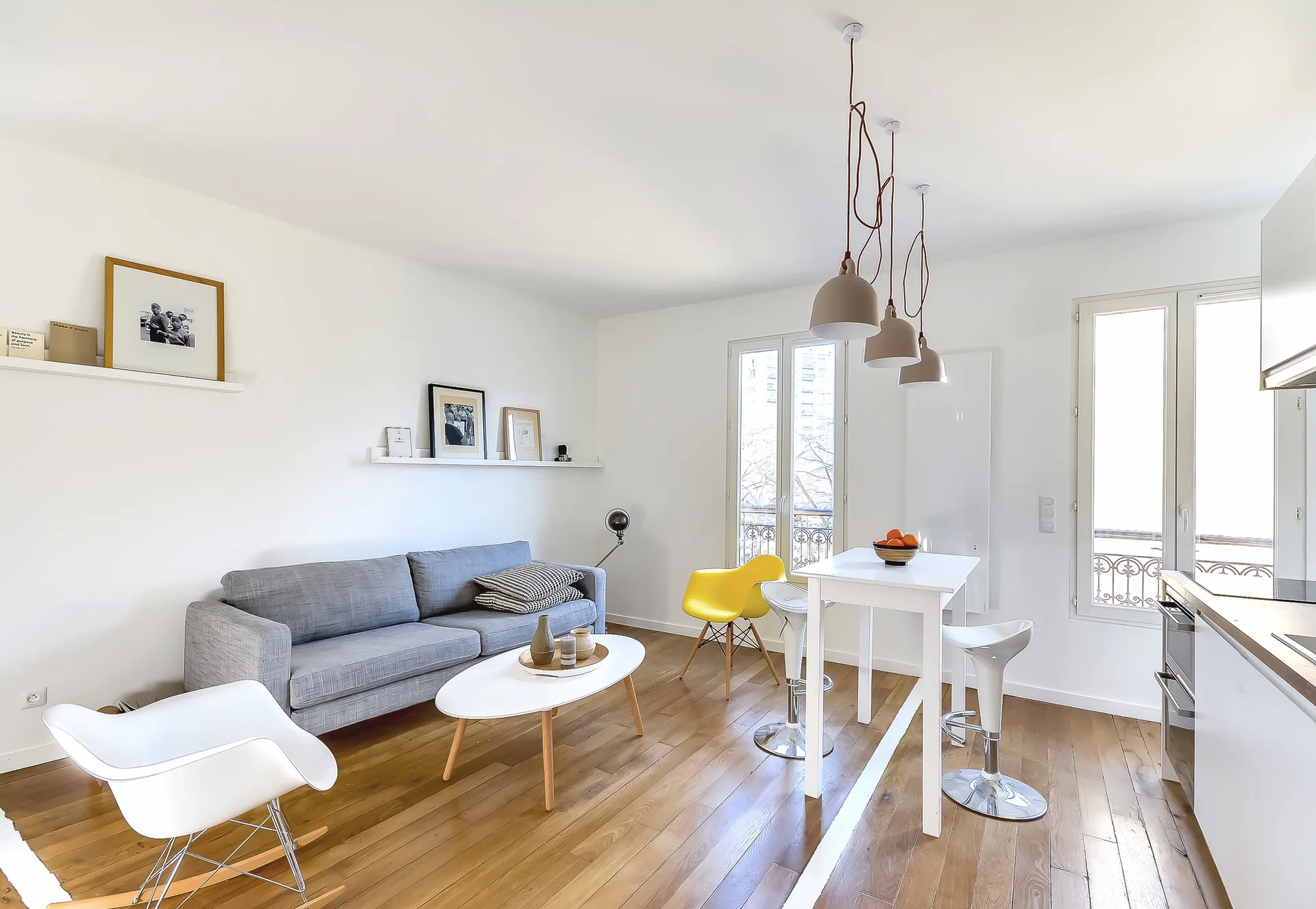 A bright open-plan apartment featuring a grey sofa, white floating art shelves, and a high-top dining table beneath three hanging pendant lights.