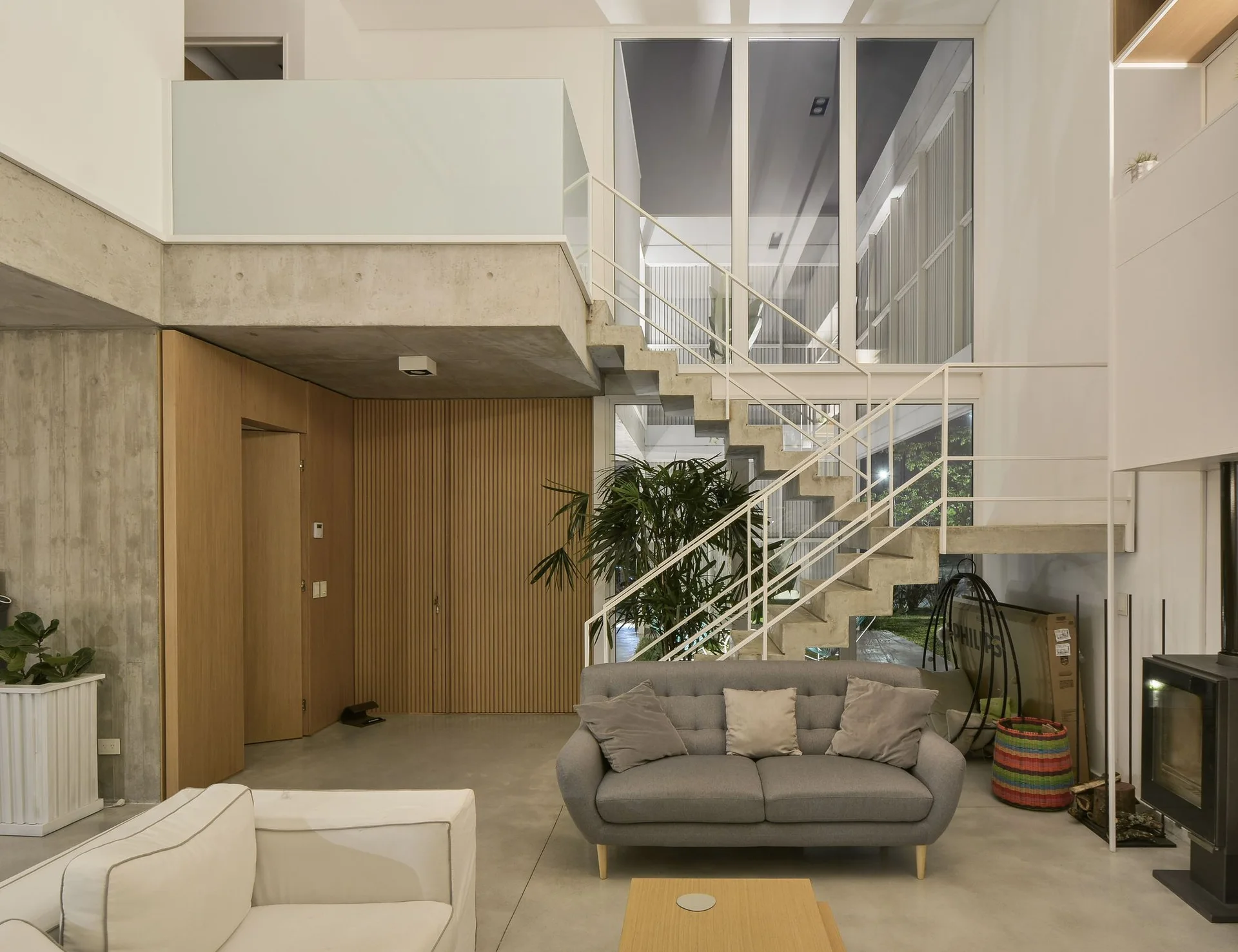 A modern double-height living room featuring an exposed concrete staircase, a grey sofa, floor-to-ceiling windows, and vertical wood slat paneling.