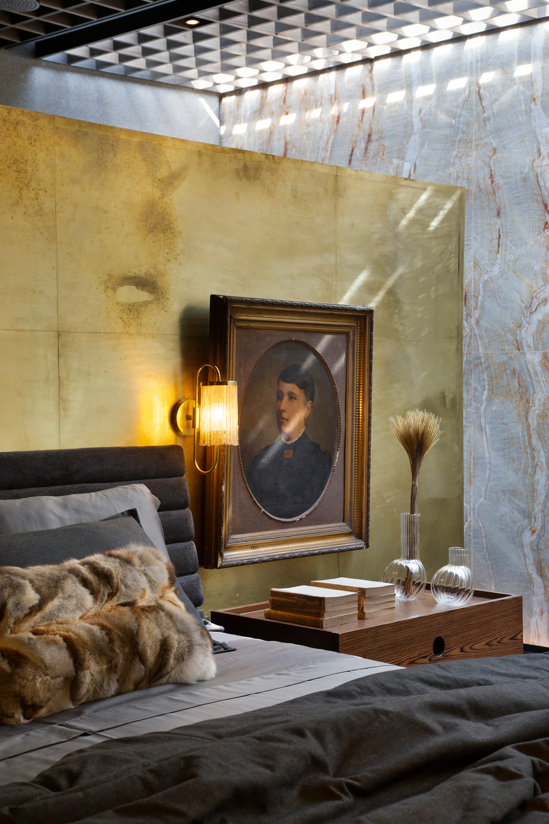 A bedroom corner features a wooden nightstand, grey bedding, a warm brass wall sconce, and a vintage framed portrait over a brass accent wall.