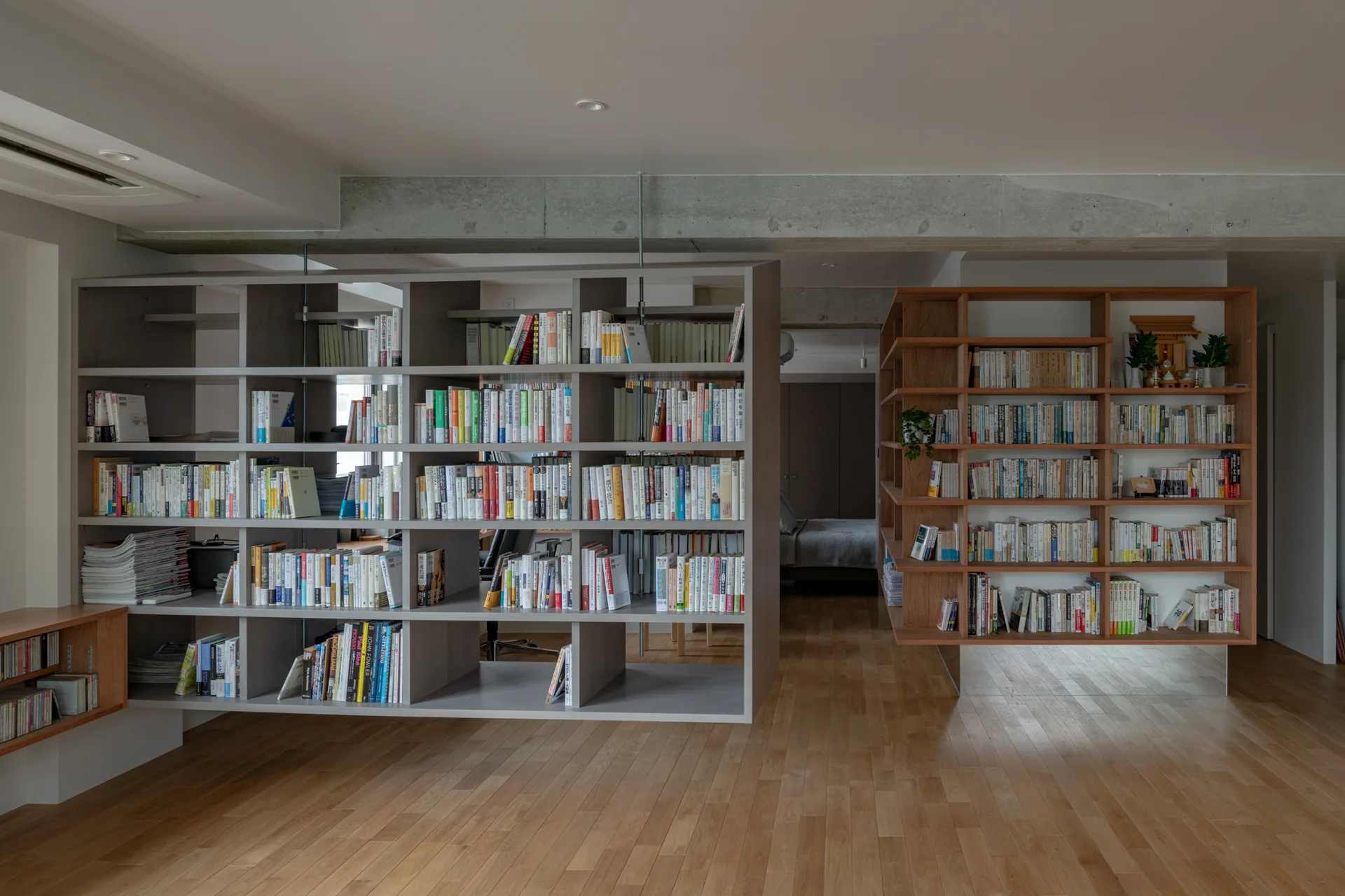 A minimalist room features a large double-sided grey bookshelf, a wood-toned floating bookshelf, light wood floors, and an exposed concrete beam overhead.