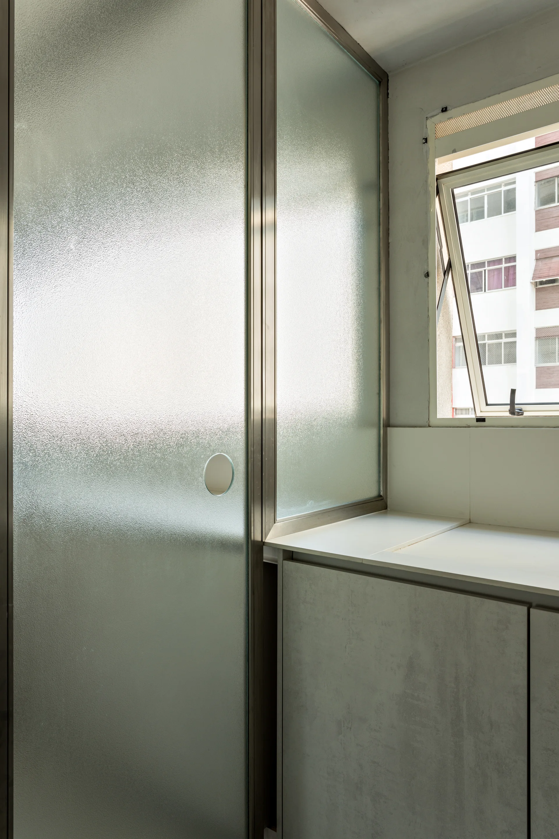 A frosted glass partition with a metal frame stands next to a grey vanity cabinet under an open casement window.