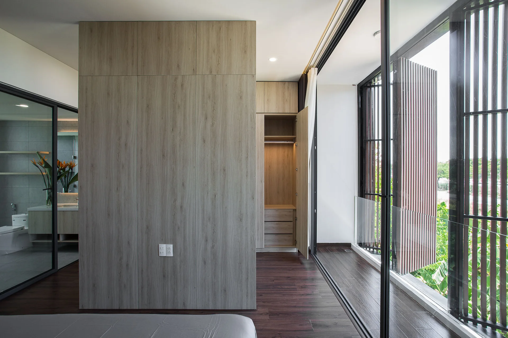 A modern bedroom features a light wood partition wall, a built-in wardrobe with interior lighting, and large sliding glass doors opening to a balcony.