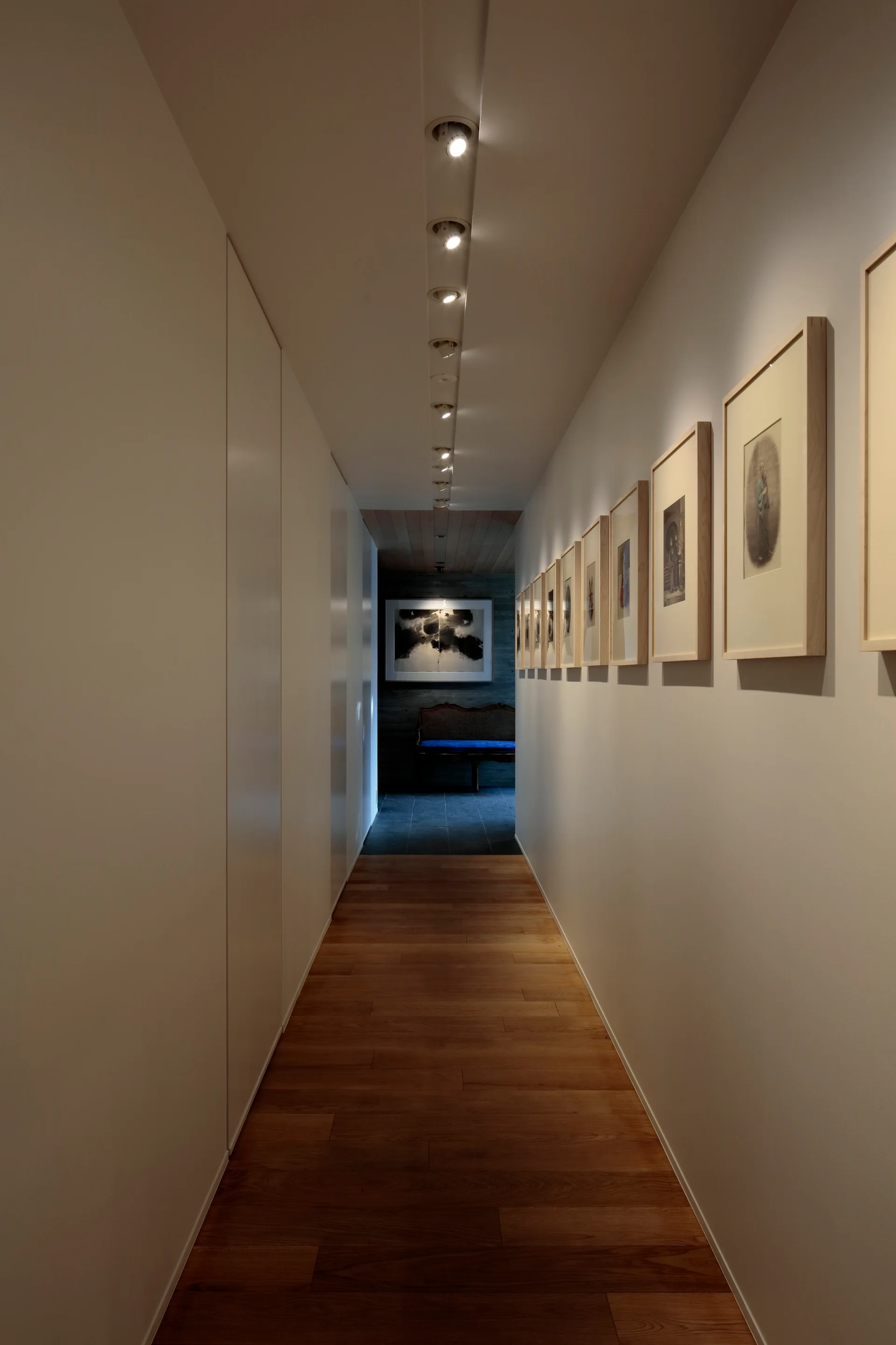 A long hallway with hardwood floors features a gallery wall of framed art illuminated by a row of recessed ceiling spotlights.