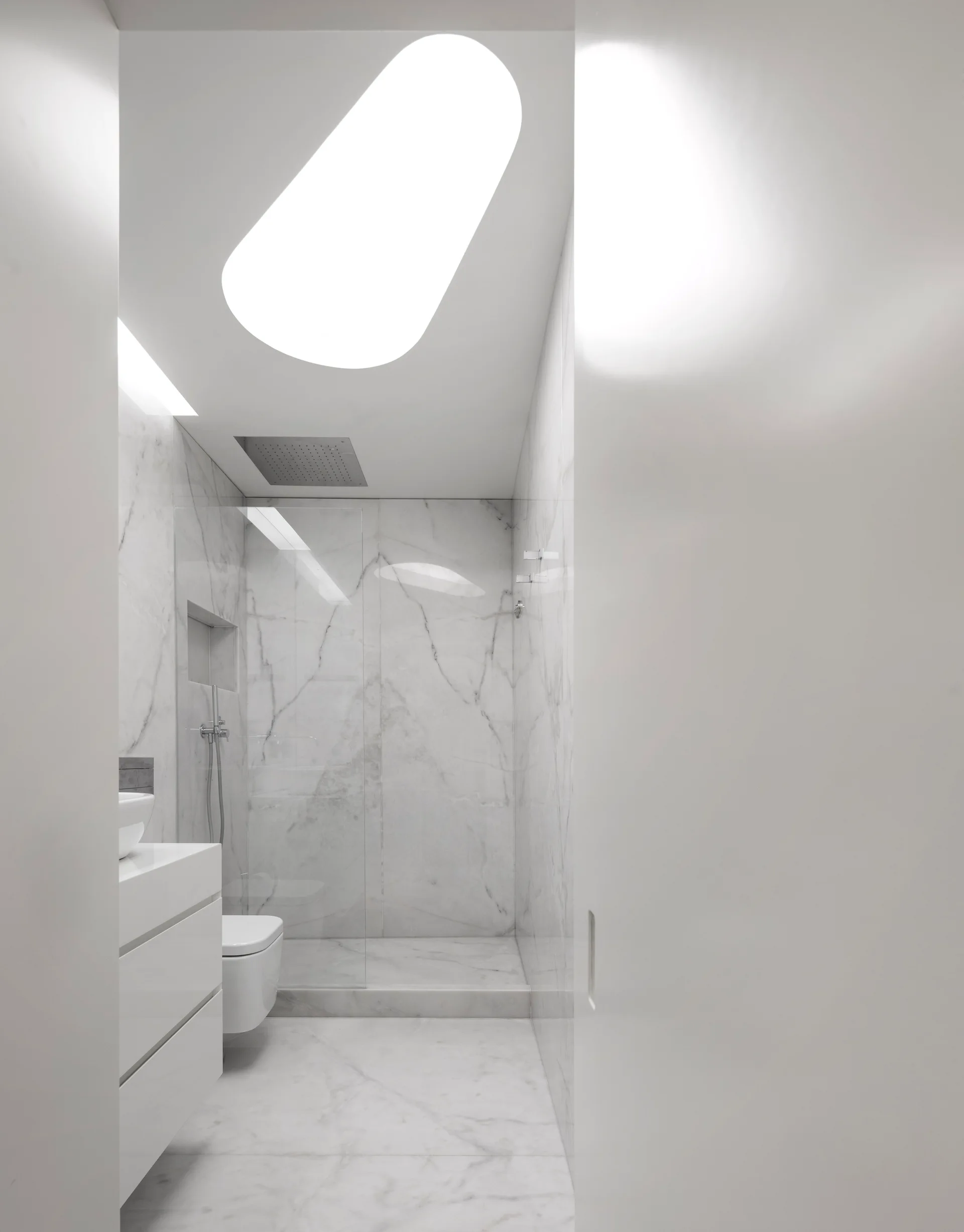 A narrow minimalist bathroom featuring white marble tile walls and floor, a glossy floating vanity, a wall-mounted toilet, and an oblong skylight.