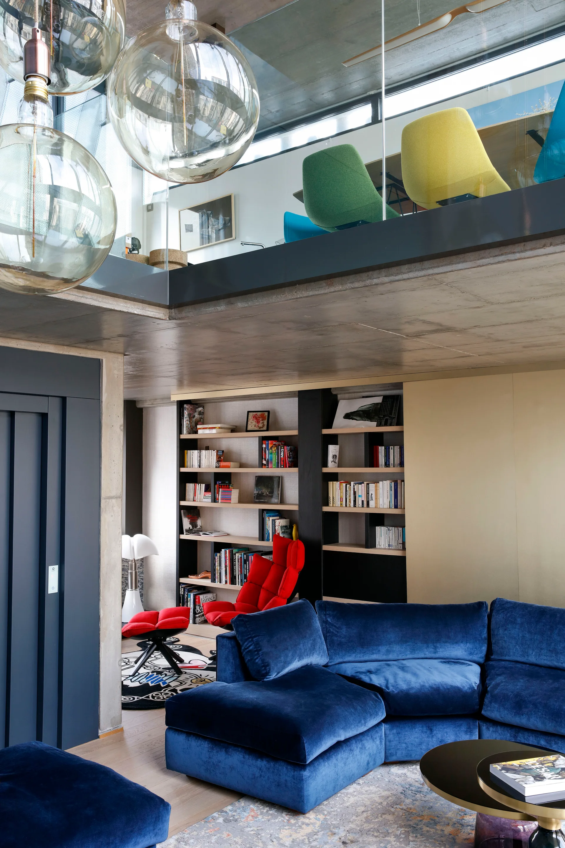 A modern loft living room features a blue velvet sectional sofa and a red lounge chair beneath a concrete ceiling and mezzanine.