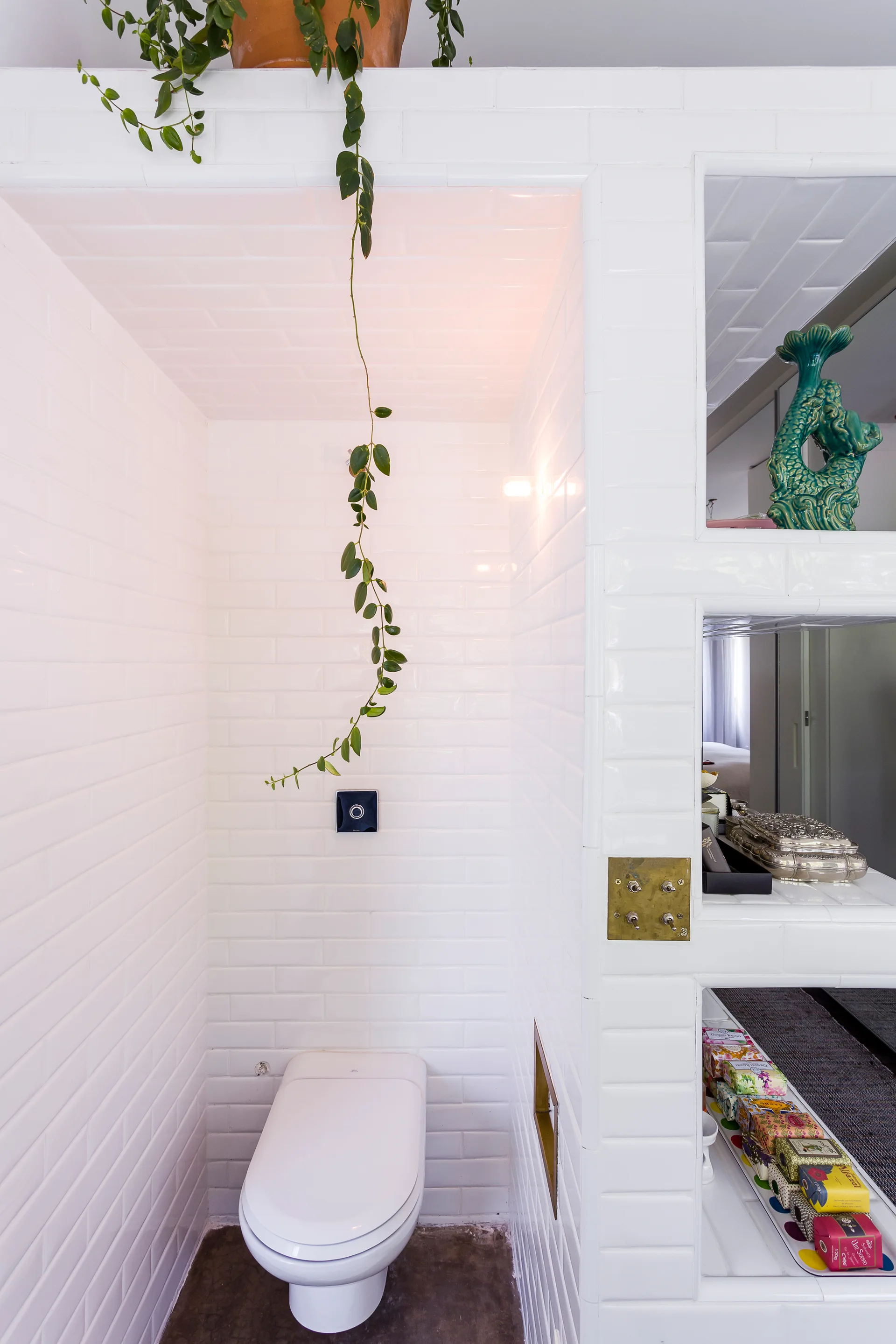 A toilet alcove clad in white subway tile features a trailing plant, built-in tiled shelves with decorative items, and a brass toggle switch.