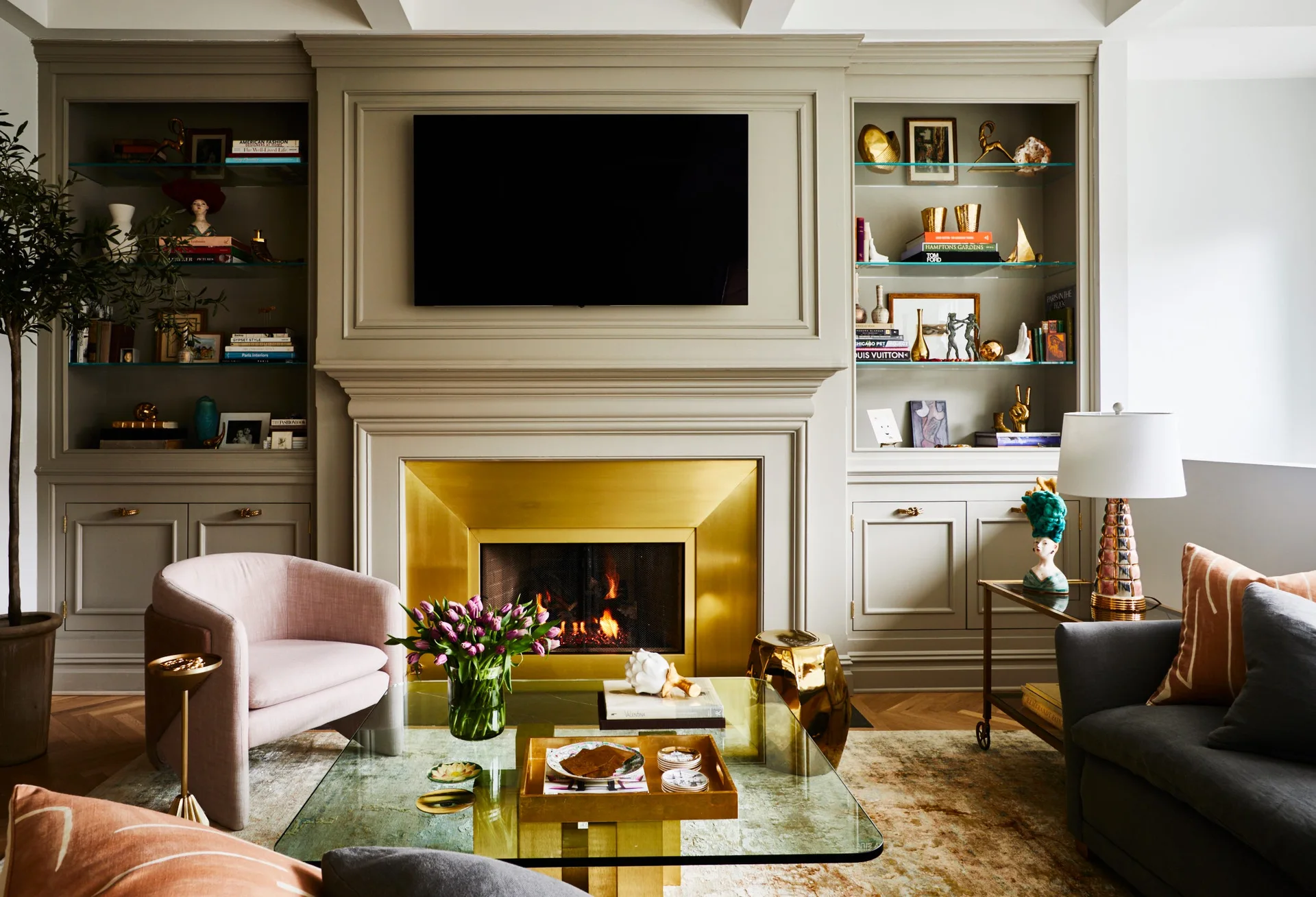 A classic living room features a brass fireplace surround flanked by taupe built-ins, with a blush pink armchair and glass coffee table.