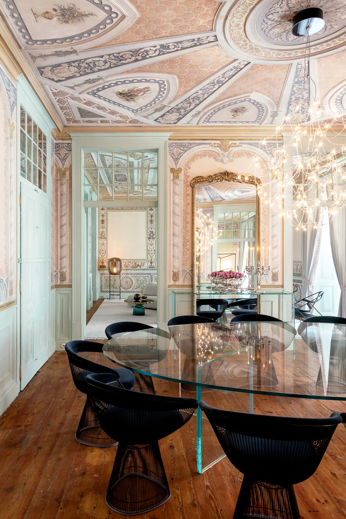 A dining room featuring an oval glass table and black wire chairs set against walls and a ceiling adorned with neoclassical fresco murals.