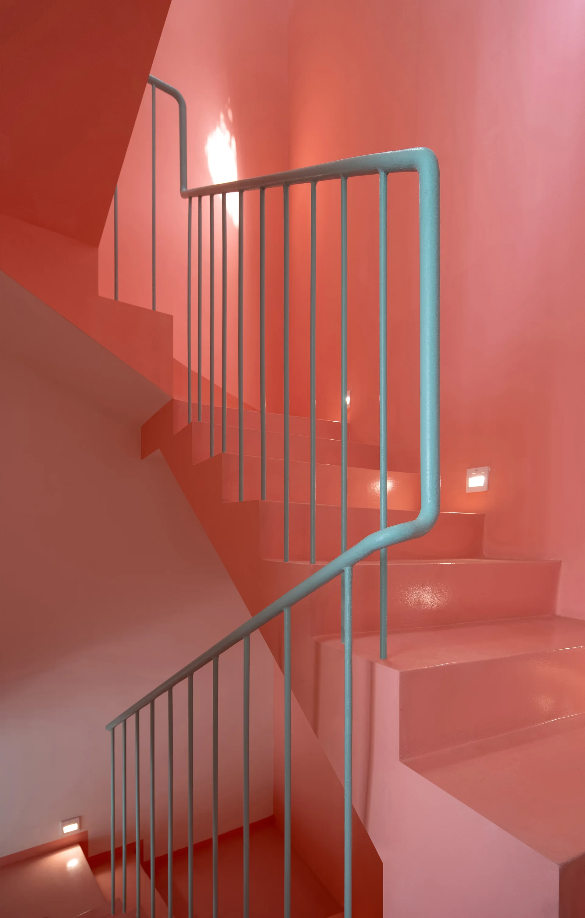 A minimalist staircase painted entirely in salmon pink features a contrasting pale teal metal handrail and small square step lights built into the wall.