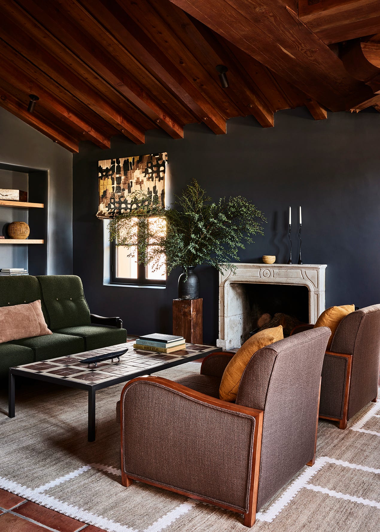 A rustic living room with dark charcoal walls, exposed wood ceiling beams, a stone fireplace, a green sofa, and brown armchairs.