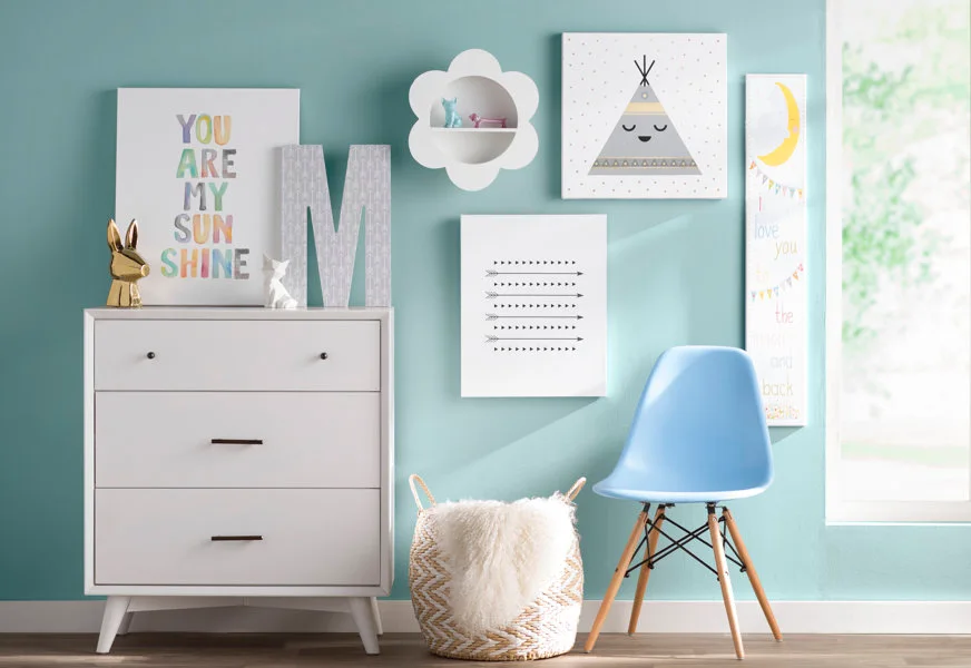 A white dresser, a blue molded chair, and a woven basket sit against a bright teal wall decorated with playful kids' wall art.