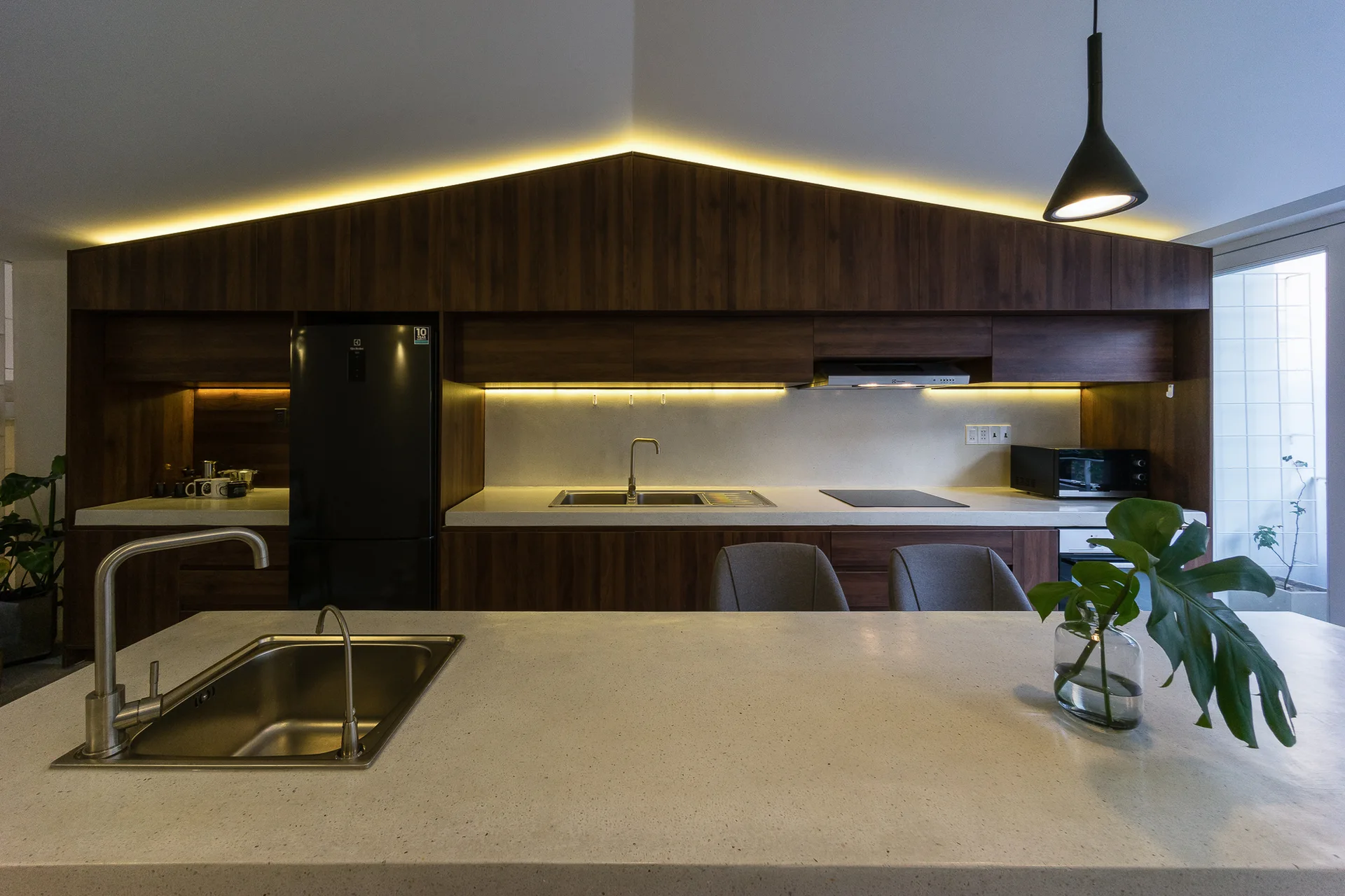 A modern kitchen features a light stone island, dark wood cabinets, integrated appliances, and warm cove lighting beneath a gabled ceiling.