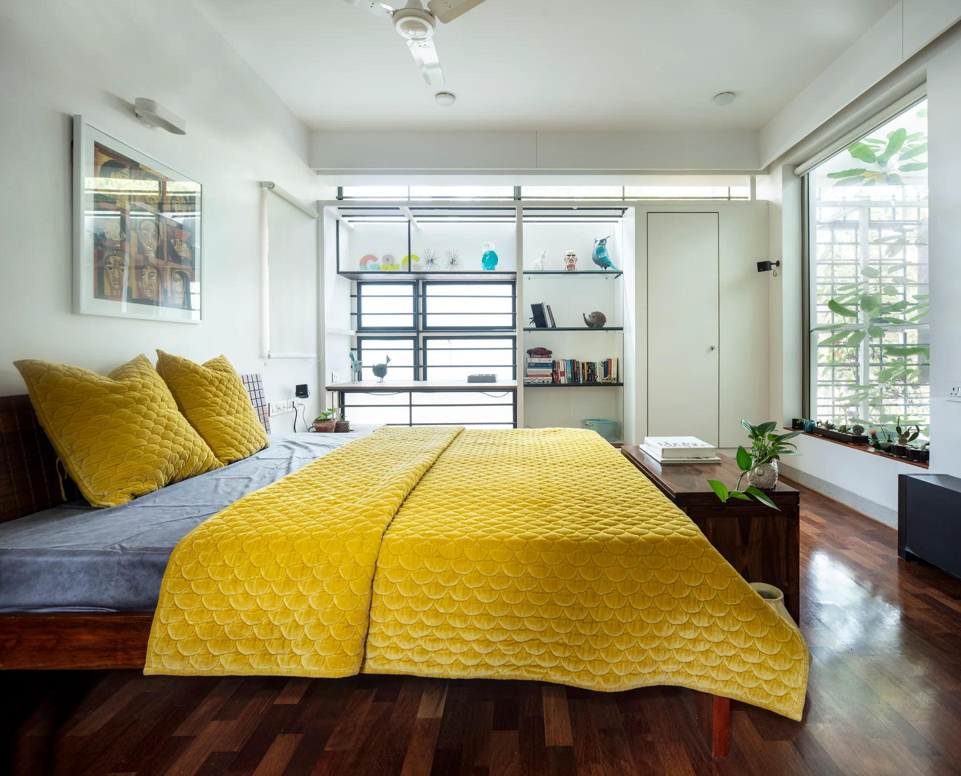 A contemporary bedroom features a wooden bed with mustard yellow bedding, dark hardwood floors, and built-in shelving framing a bright window.