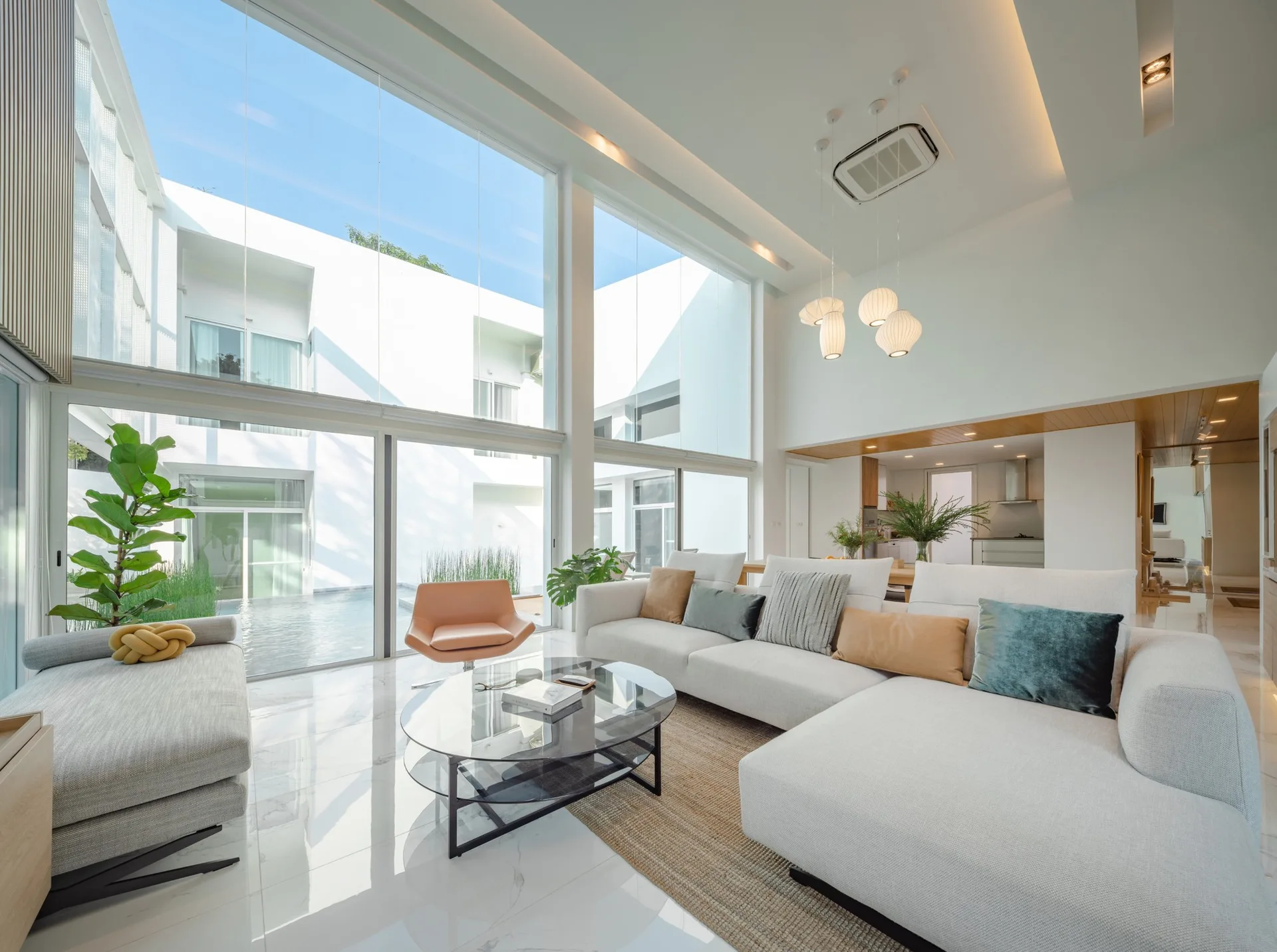 A double-height living room featuring a white sectional sofa, glass coffee table, and massive windows overlooking an outdoor courtyard.