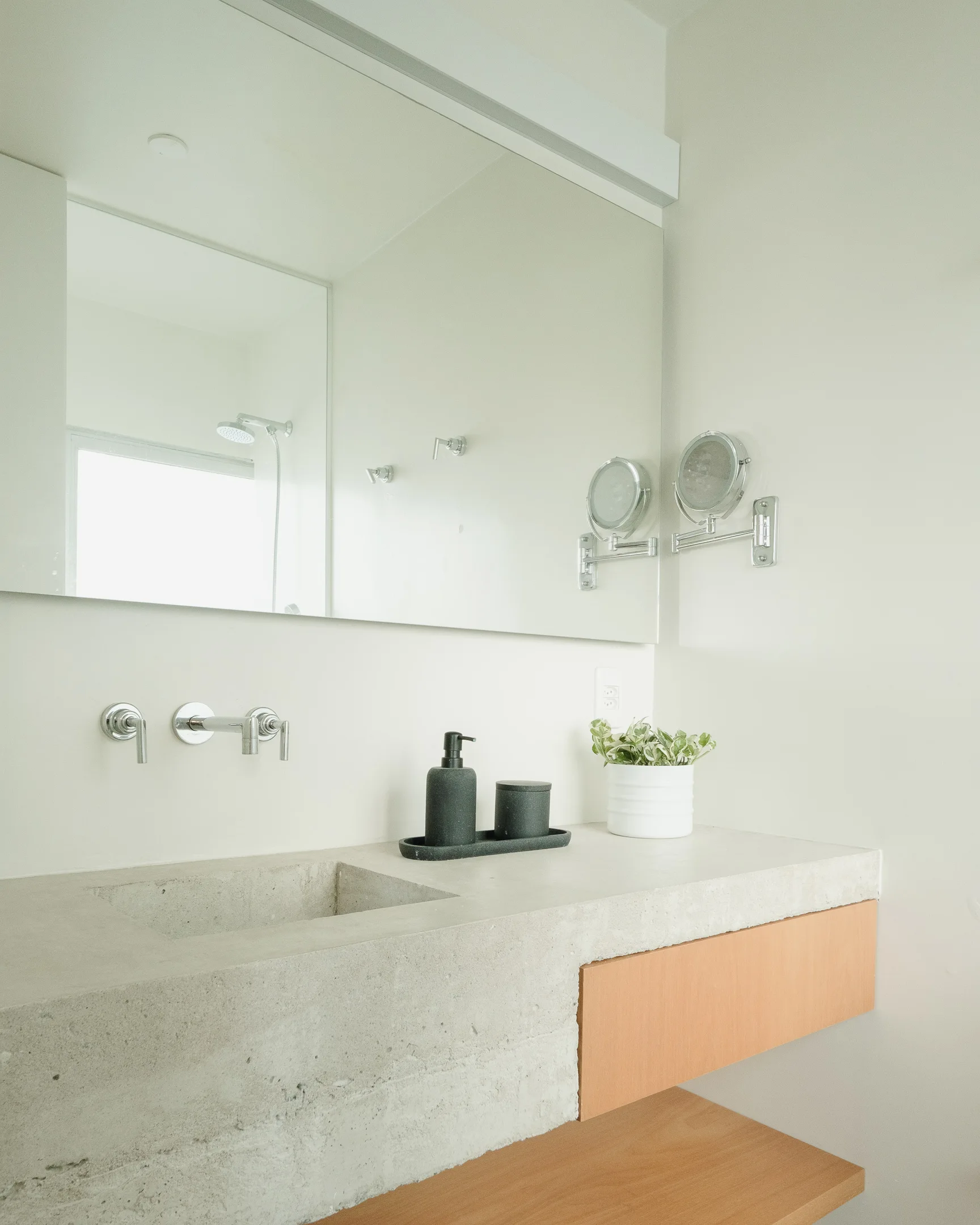 A modern bathroom vanity featuring an integrated concrete sink, light wood cabinetry, a large wall mirror, and chrome wall-mounted faucets.