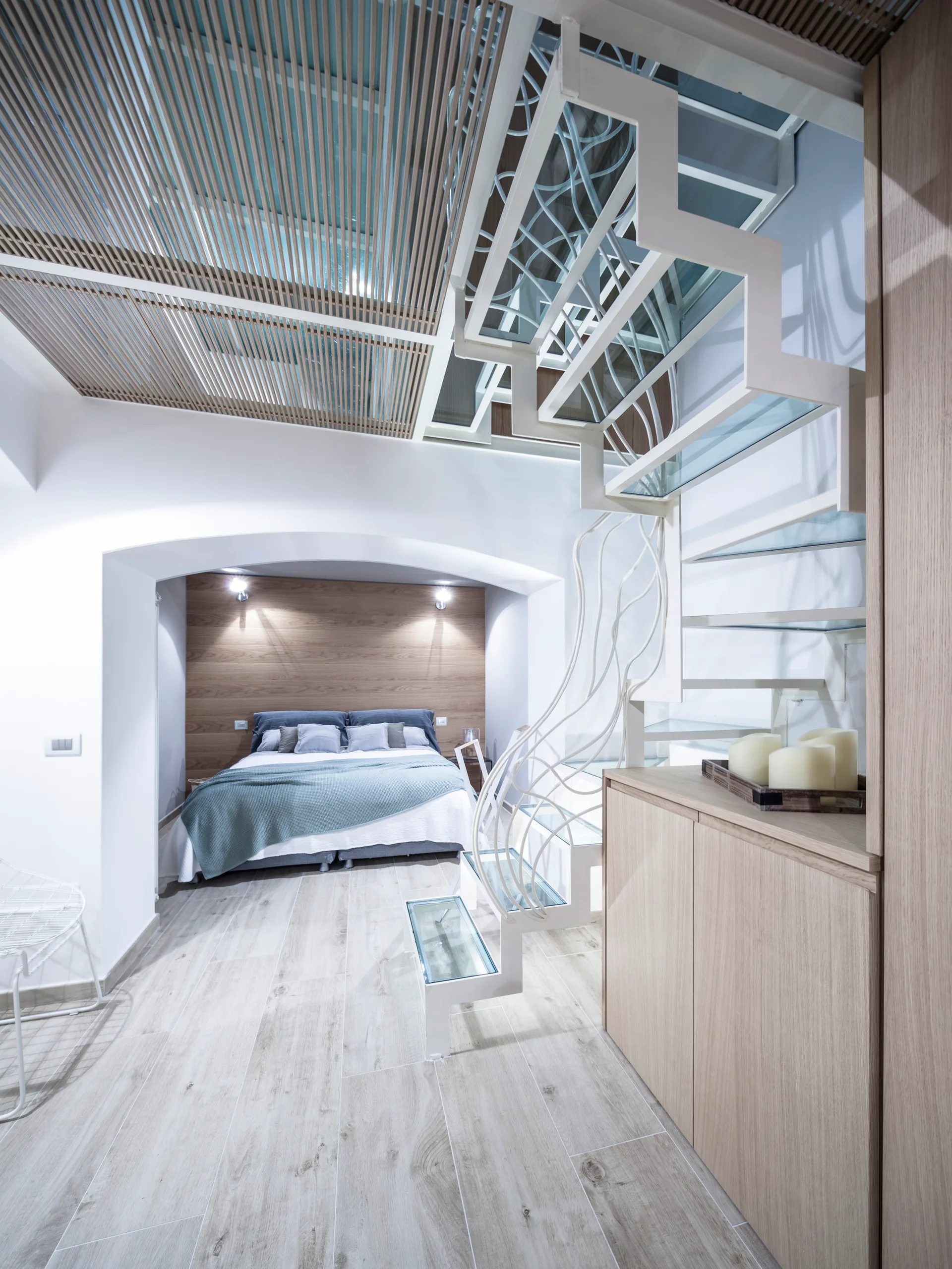 A contemporary bedroom featuring a white metal staircase with glass treads, an arched alcove framing the bed, and a wood-slatted ceiling.