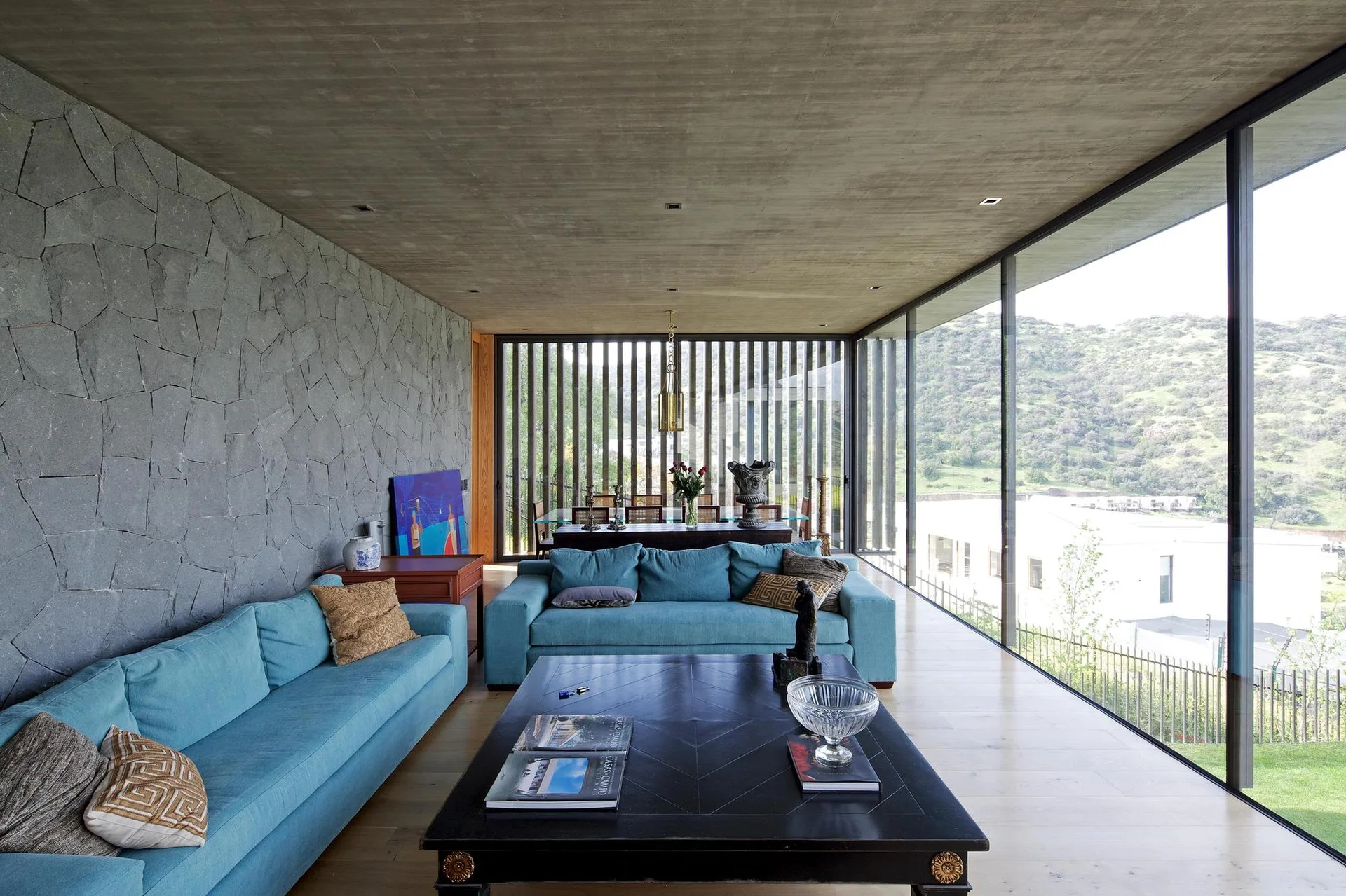 A spacious modern living room features two teal sofas, a dark wood coffee table, a textured grey stone wall, and floor-to-ceiling glass windows.