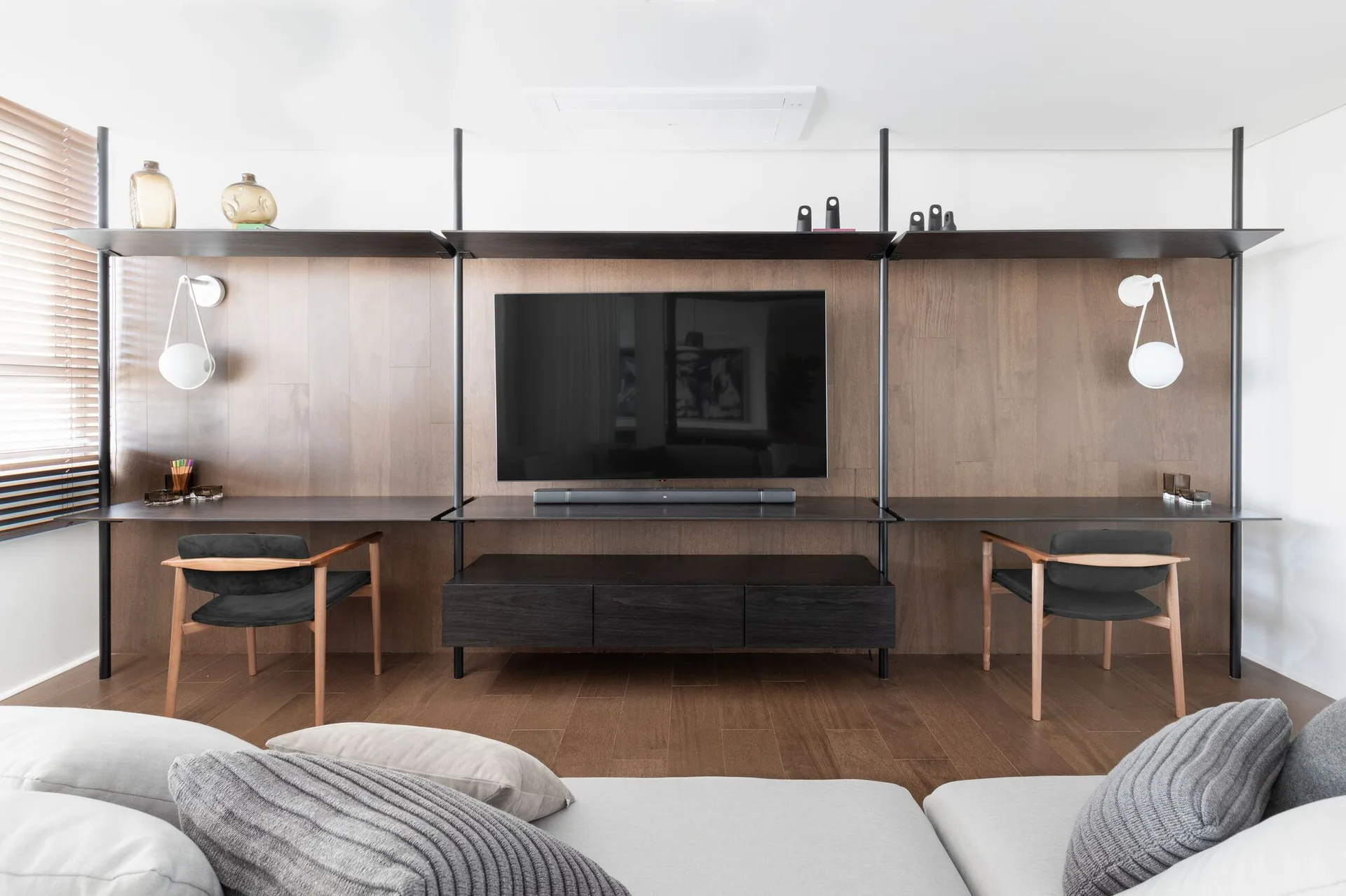 A modern living room features a wood-paneled wall with an integrated media unit, two desks with chairs, and a grey sofa in the foreground.