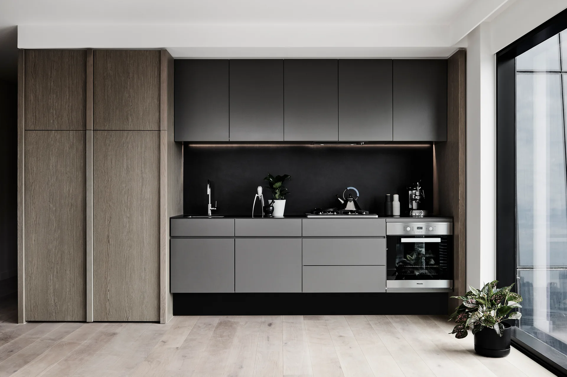 A minimalist kitchen featuring matte grey cabinetry, a wood-grain pantry cabinet, light wood floors, and integrated under-cabinet lighting.
