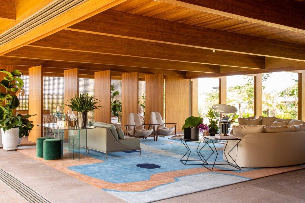 A modern open-plan living room features exposed wood ceiling beams, a blue and orange geometric rug, cream sofa, and vertical wood partition screens.