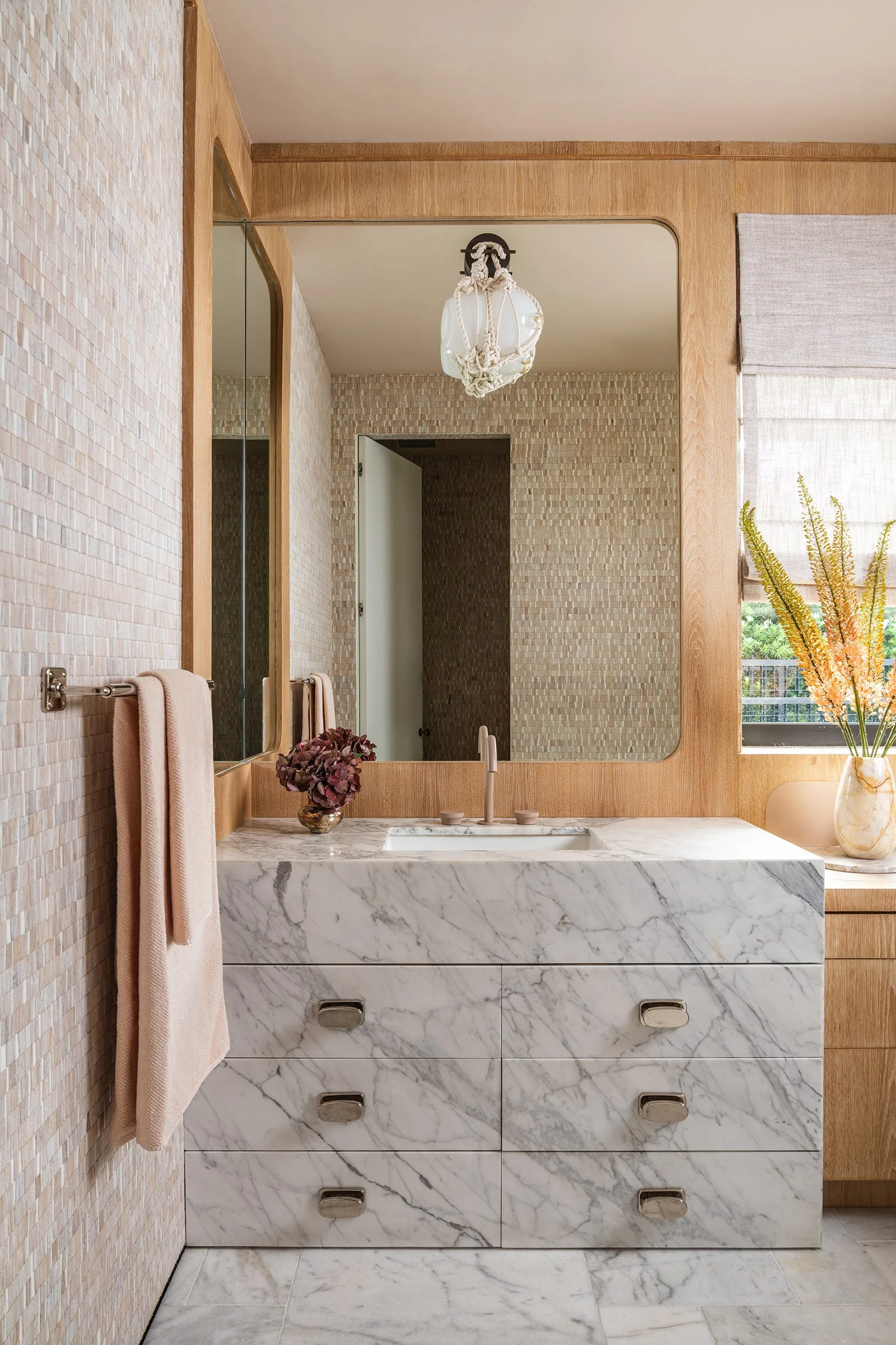 A bathroom vanity features marble drawer fronts and countertop set against small beige mosaic wall tiles under a large wood-framed mirror.