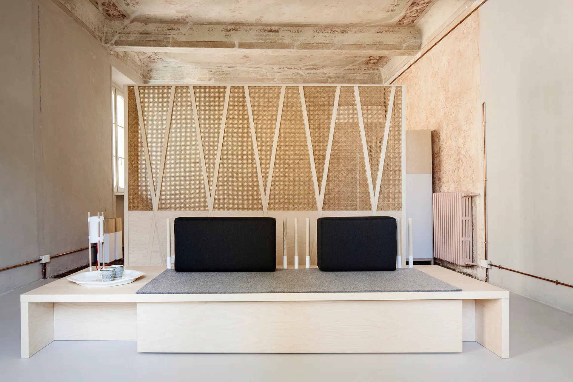 A low wooden platform daybed with black cushions sits in front of a tall geometric cane screen divider in a minimalist room with exposed ceiling beams.