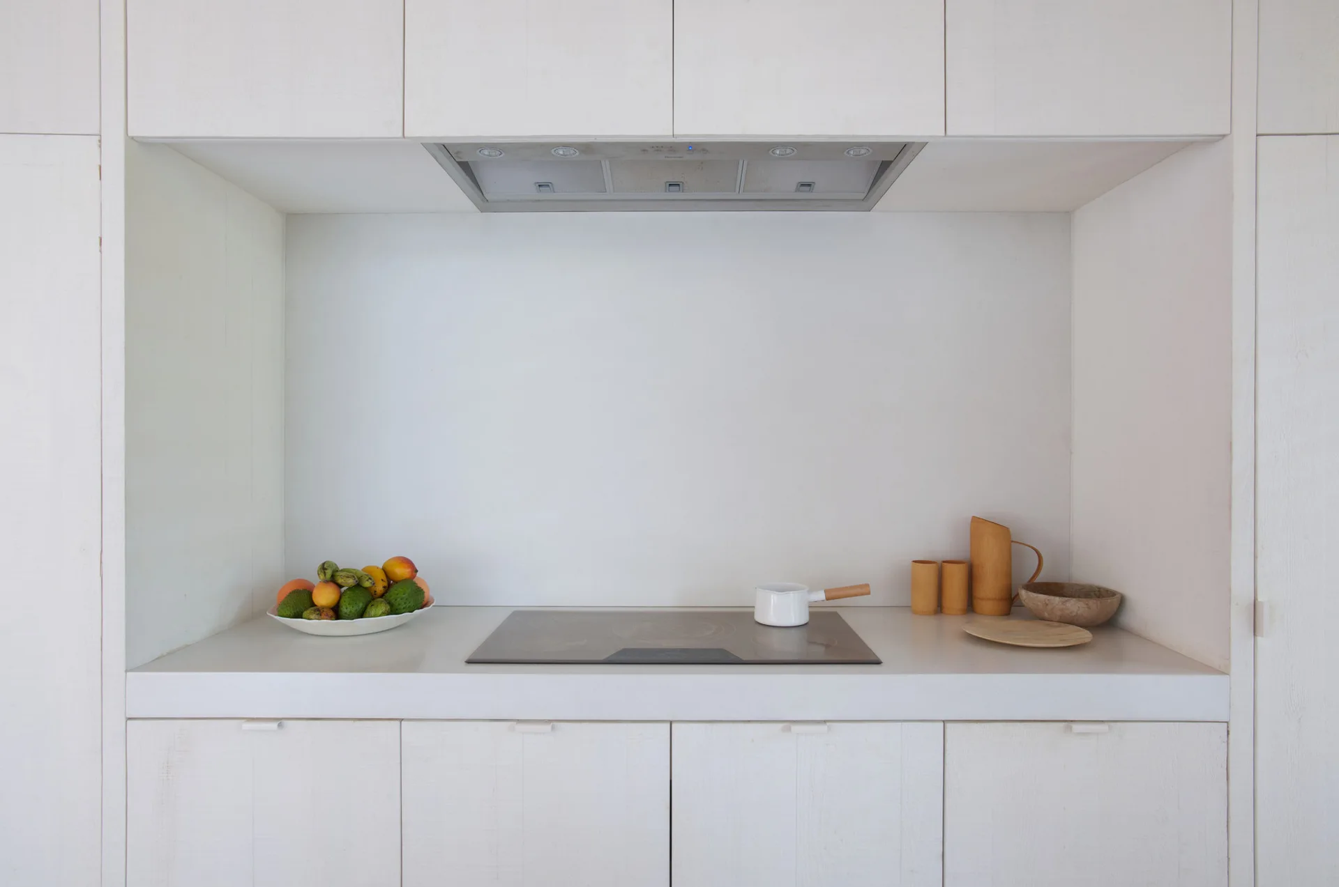 A minimalist kitchen alcove features white-washed wood cabinetry, a recessed stovetop, and simple wooden accessories.