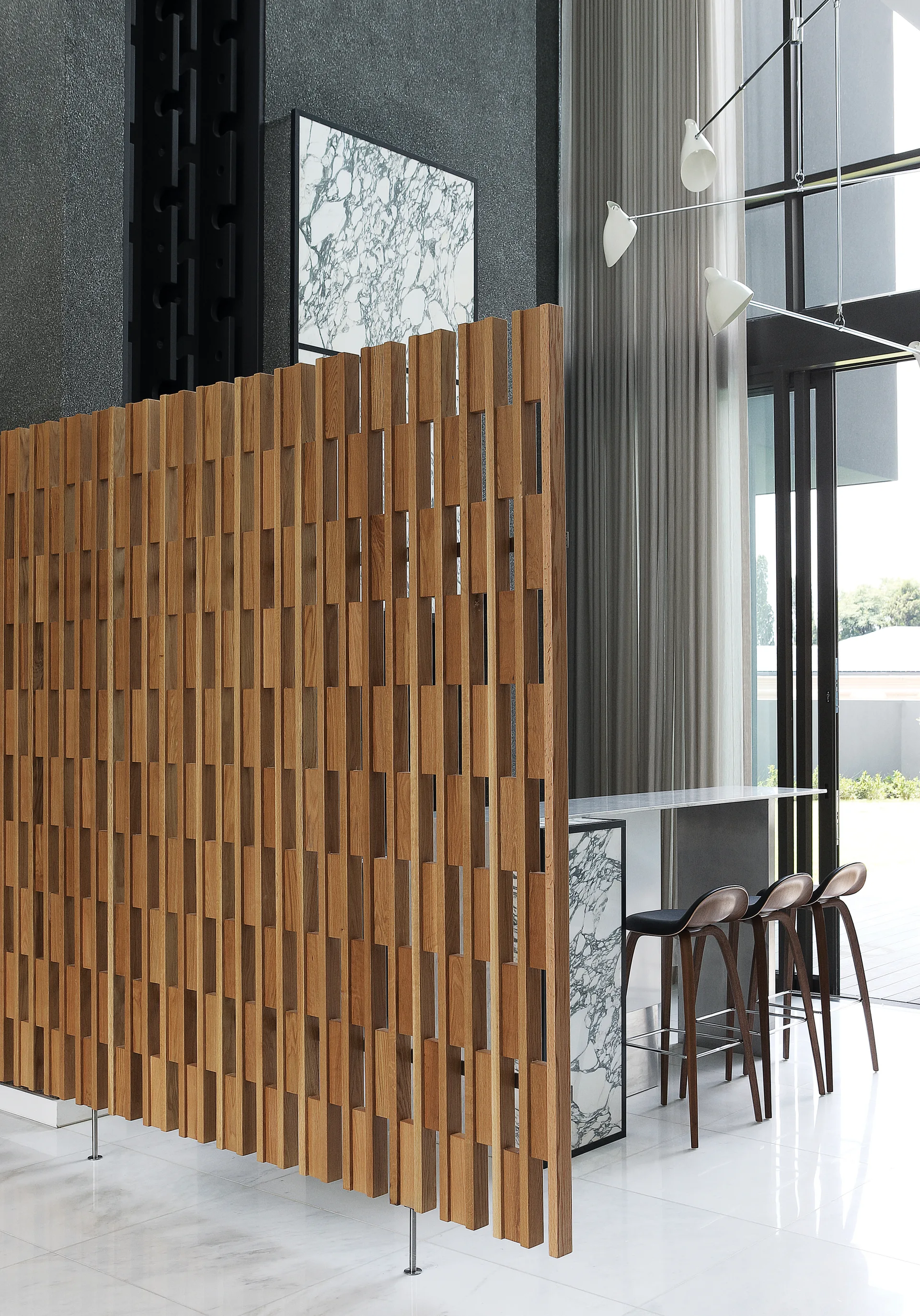 A modern kitchen bar features a slatted oak wood room divider, marble counter, curved wooden bar stools, and a branching light fixture.